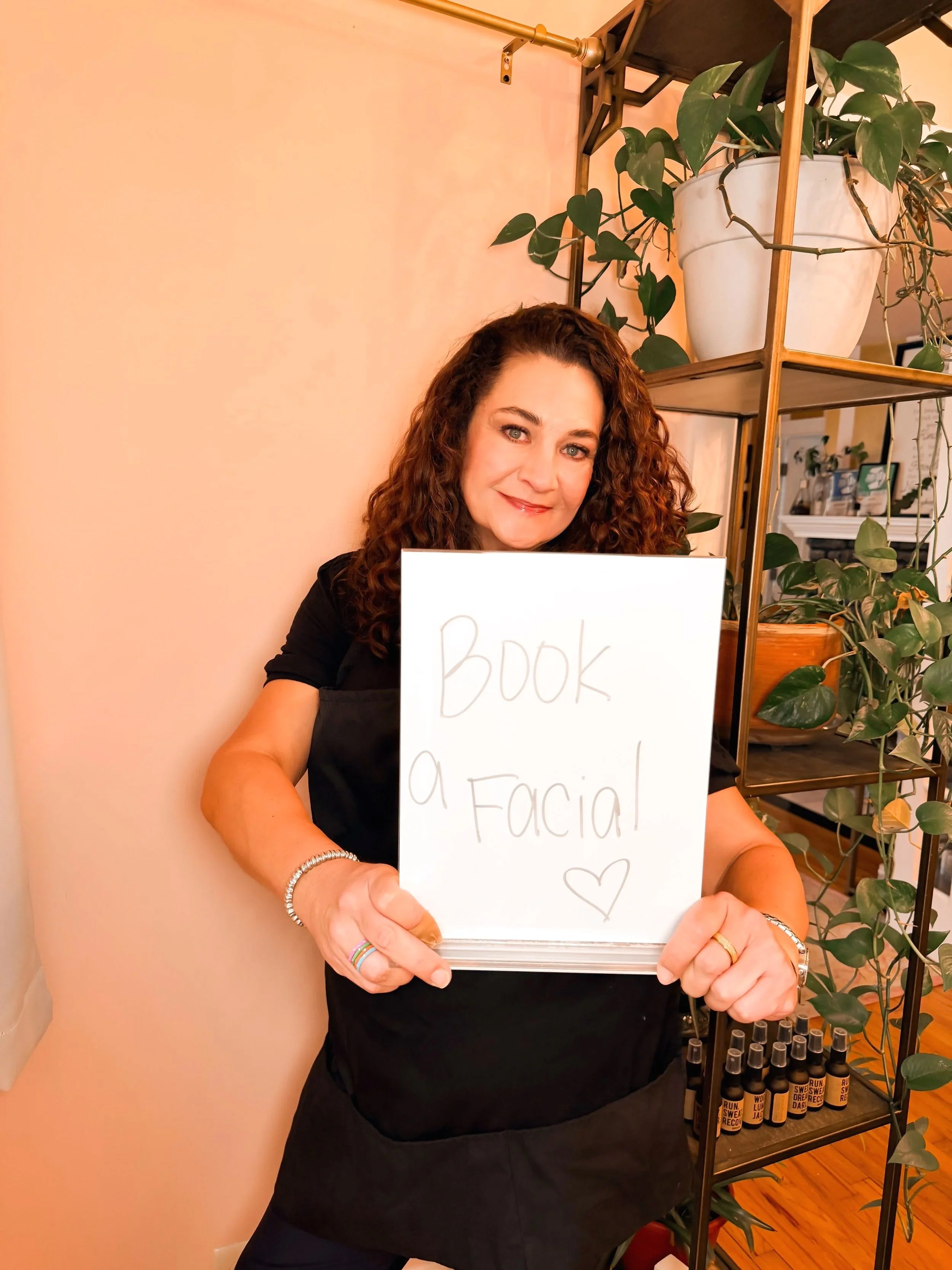 A woman with curly brown hair and light skin holding a white sign that says 'Book a Facial' with a heart drawing on it, standing next to a black metal shelving unit containing potted plants and small bottles in a cozy room with warm lighting.