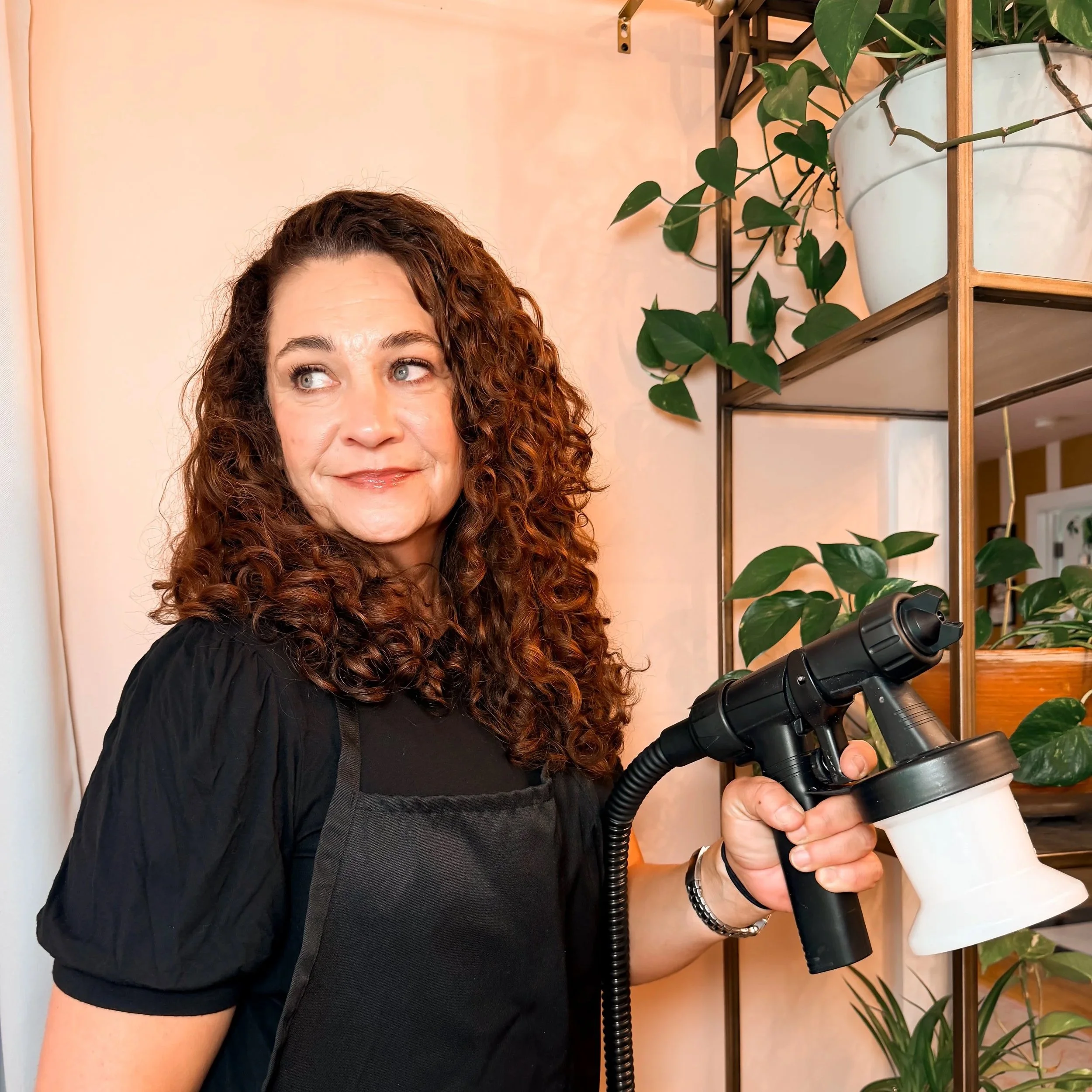 A woman with curly brown hair, wearing a black top, holding a black telescope indoors with green potted plants and shelves in the background.