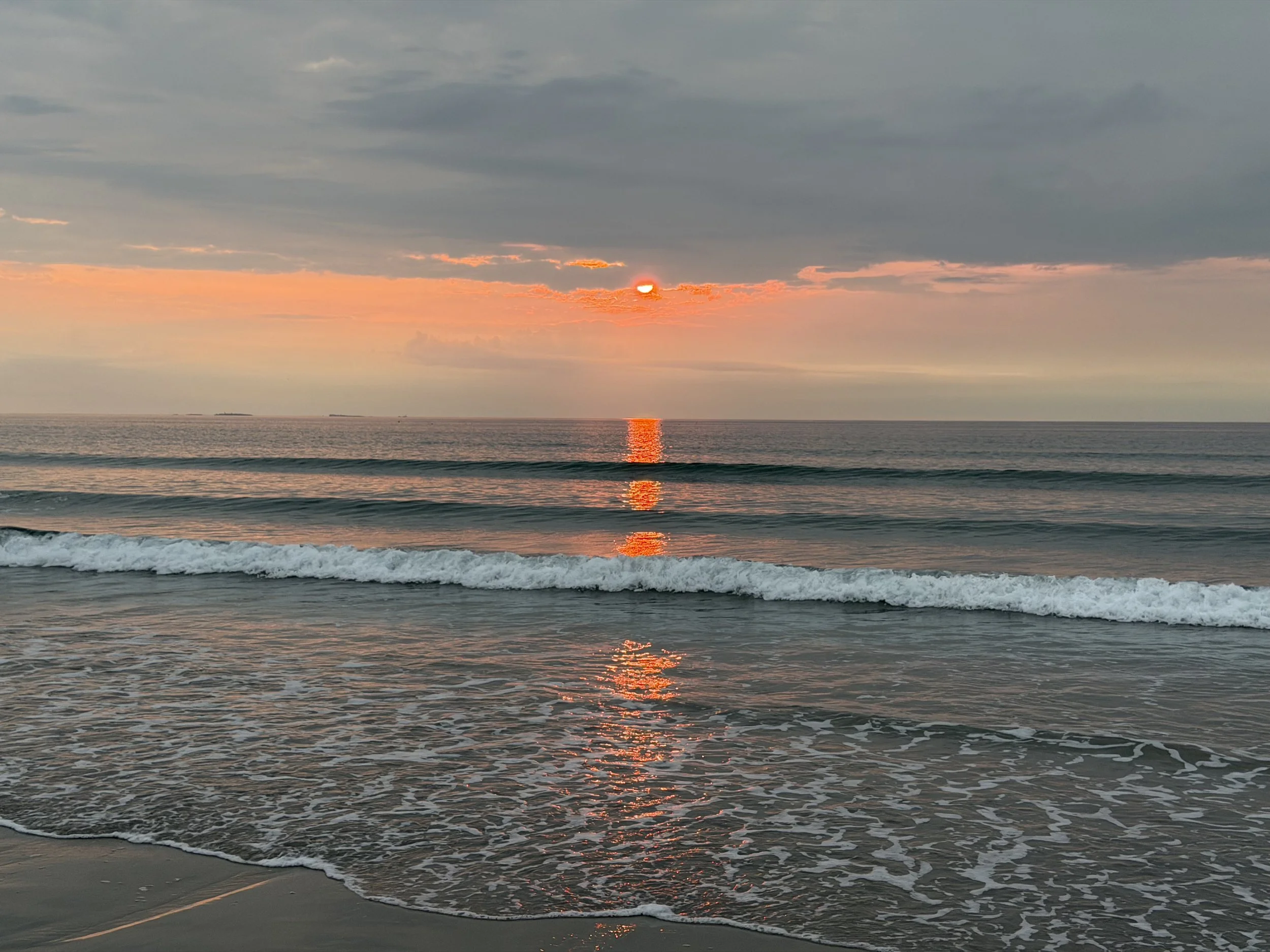 Sunset over the ocean with clouds in the sky, reflecting on the water with gentle waves.