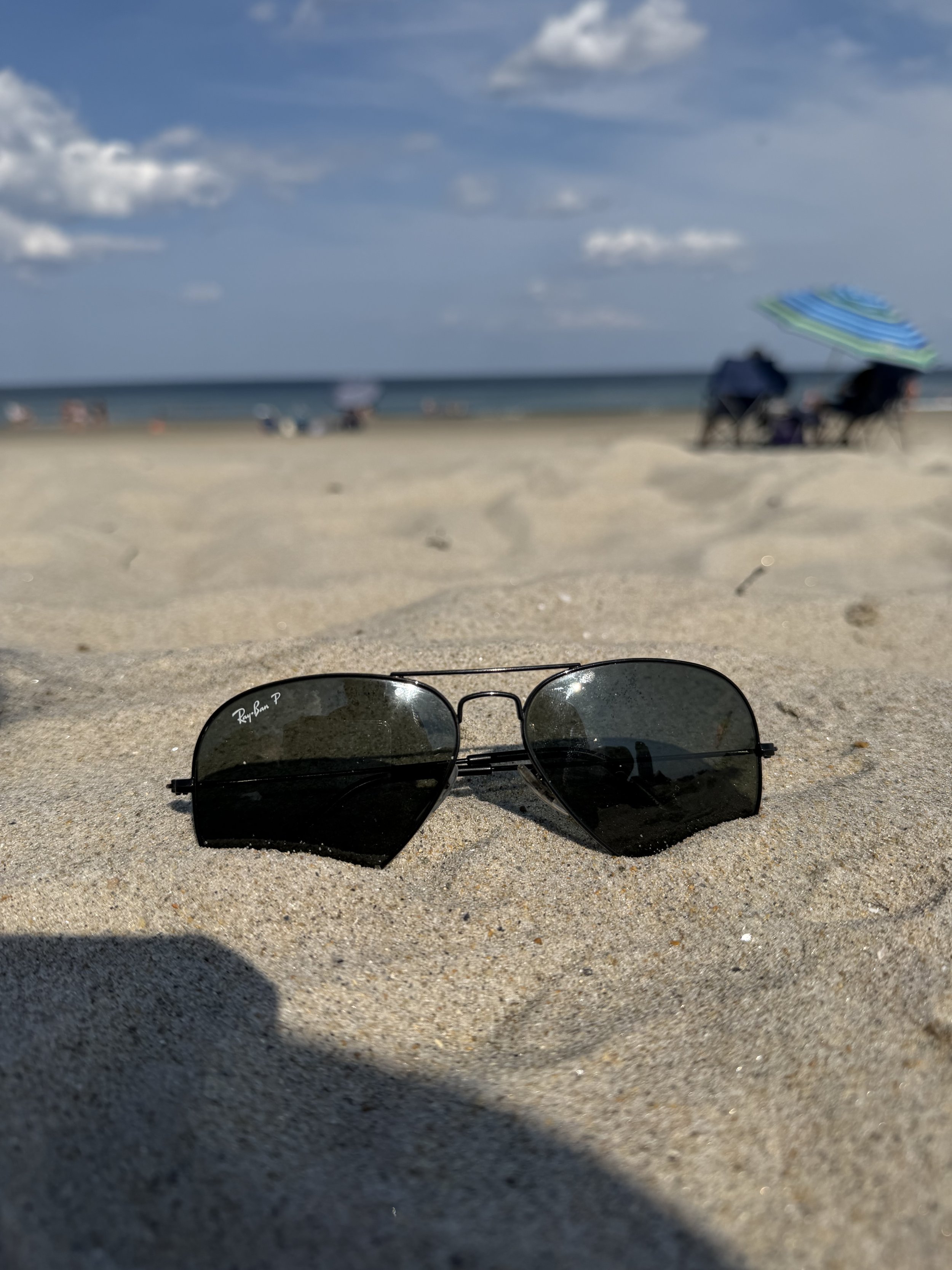 A pair of sunglasses resting on sandy beach near the shoreline, with an umbrella and beach chairs in the background under a partly cloudy sky.