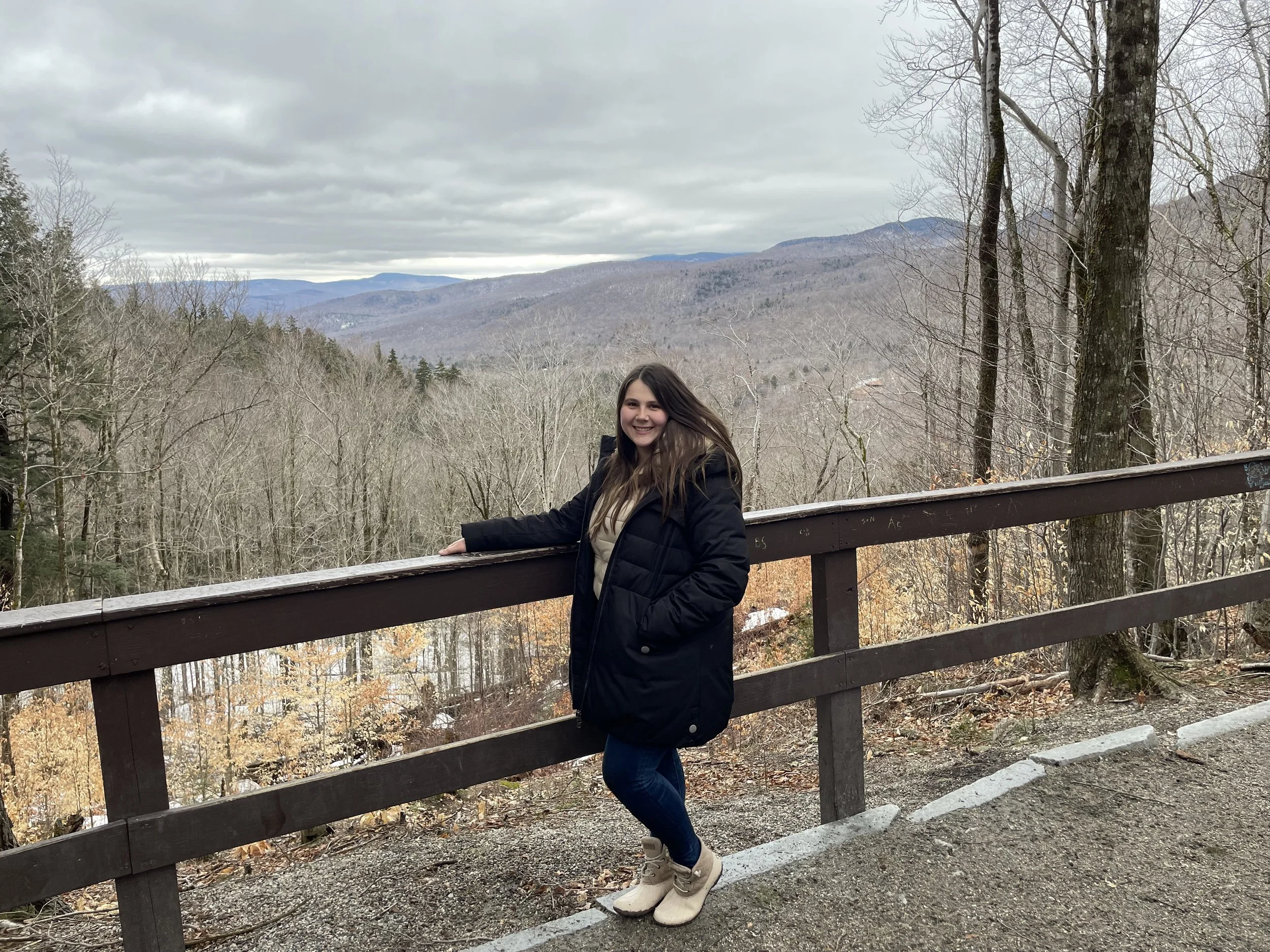 A smiling woman stands at a wooden railing with a mountain landscape and leafless trees in the background on a cloudy day.