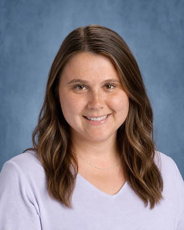 Young woman with brown hair and light skin smiling at the camera, wearing a light purple top and a delicate necklace, against a blue background.