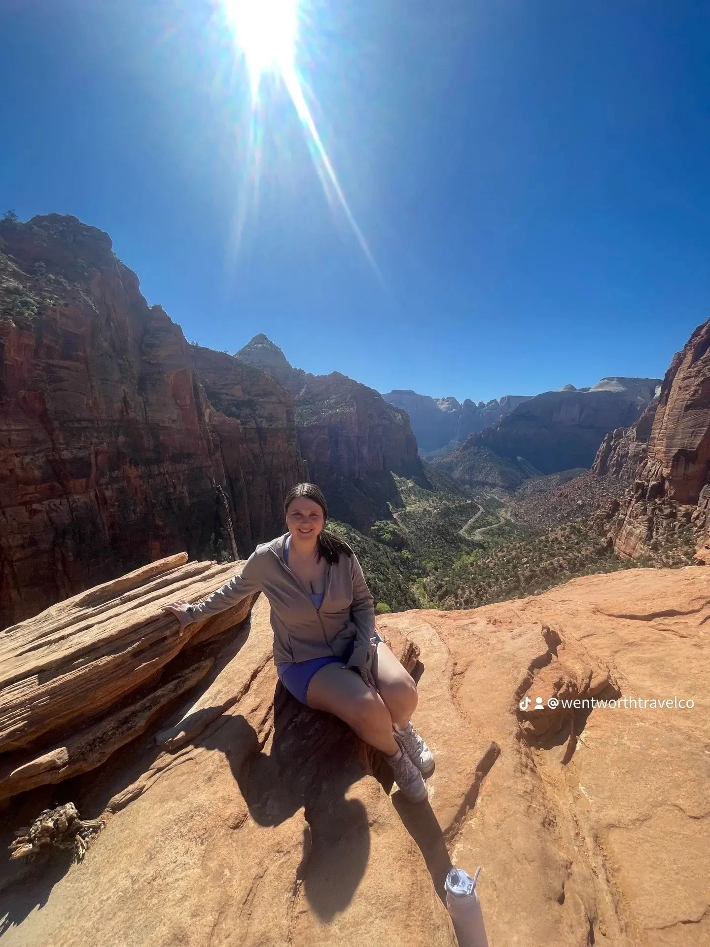 A woman sitting on a rock ledge with the Grand Canyon in the background, under a bright sun in a clear blue sky.