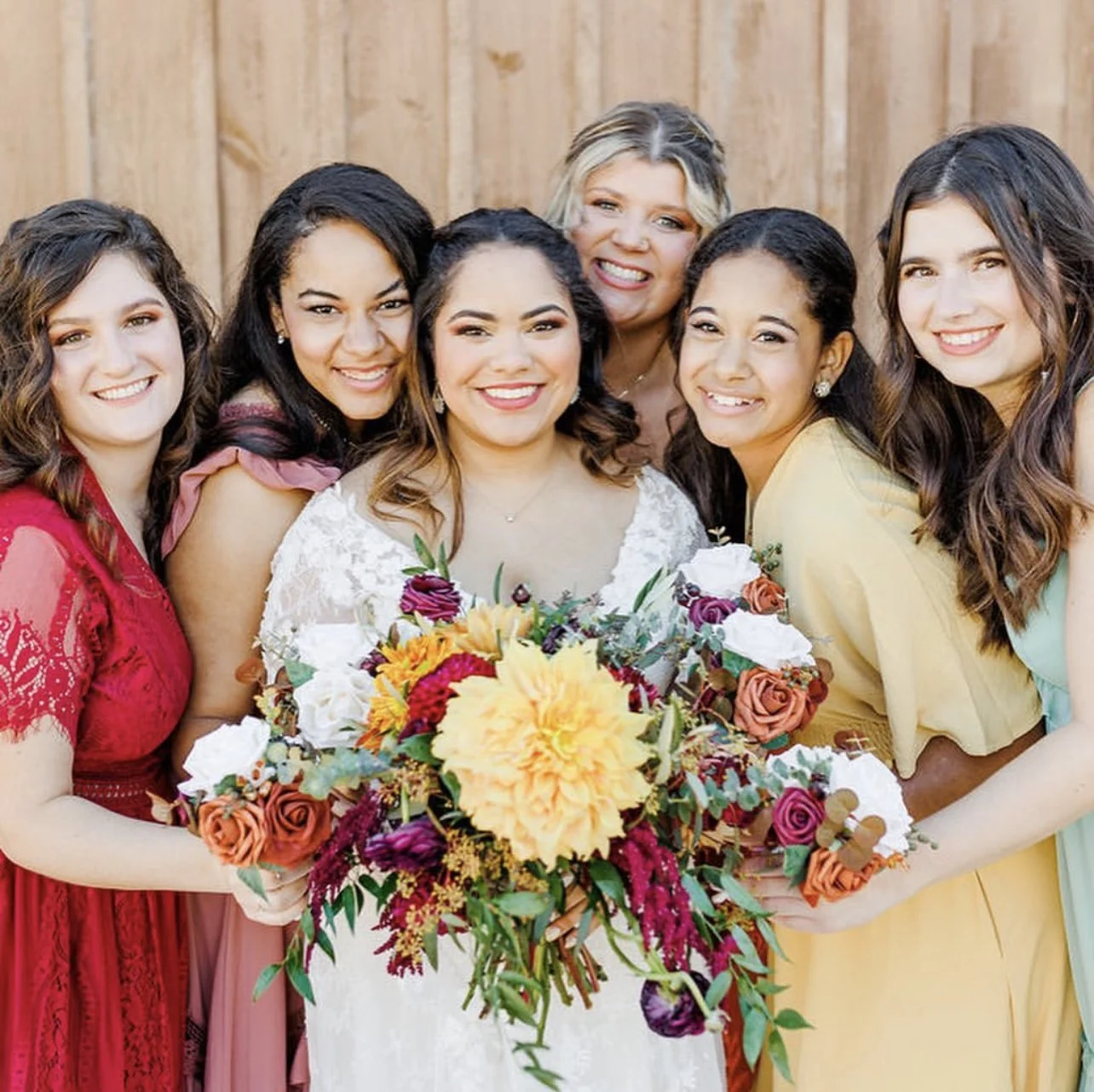 Group of seven women, including a bride holding a large bouquet, standing close together in front of a wooden fence, smiling at the camera.