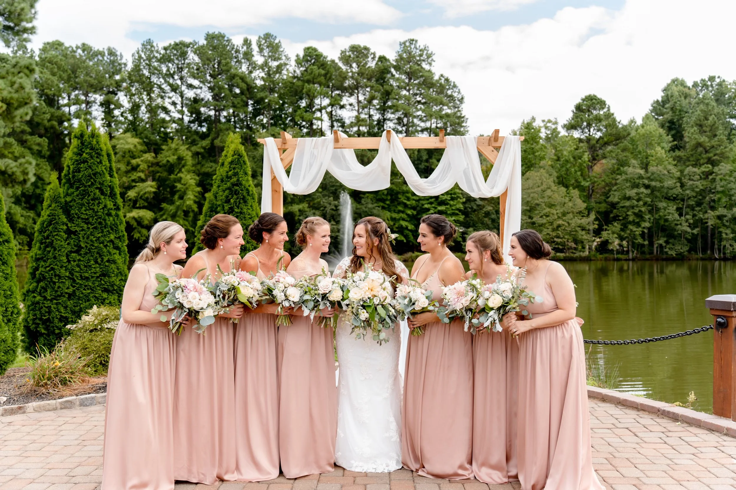 A bride and six bridesmaids standing outdoors near a pond and surrounded by green trees, with a wooden arch draped with white fabric above them, during a wedding ceremony.