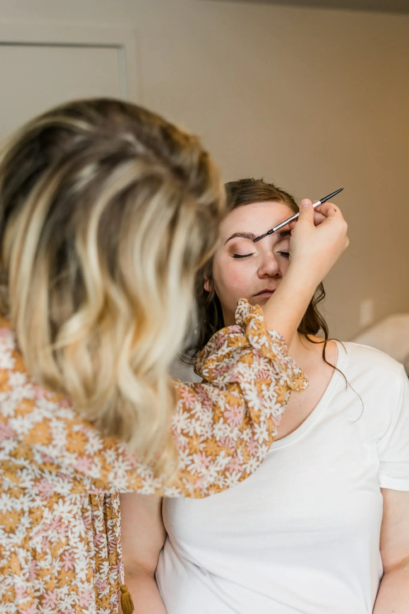 A makeup artist applying makeup to a woman's eyebrows using a brush. The woman has her eyes closed and is wearing a white top. The makeup artist is wearing a floral-patterned shirt.