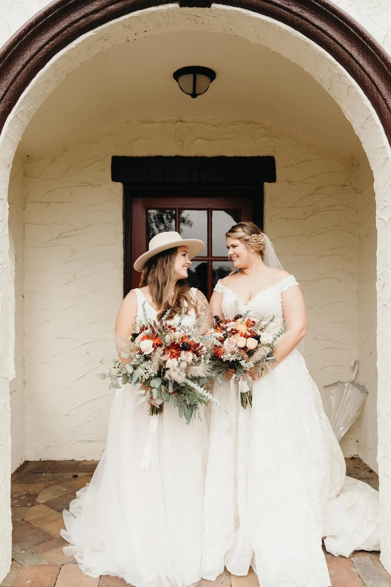 Two women in wedding dresses holding bouquets and smiling at each other under an archway.
