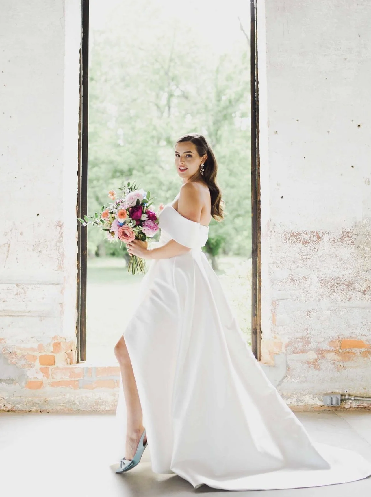 Bride in a white wedding dress standing in a rustic window frame holding a bouquet of pink, purple, and white flowers, outdoors with green trees in the background.