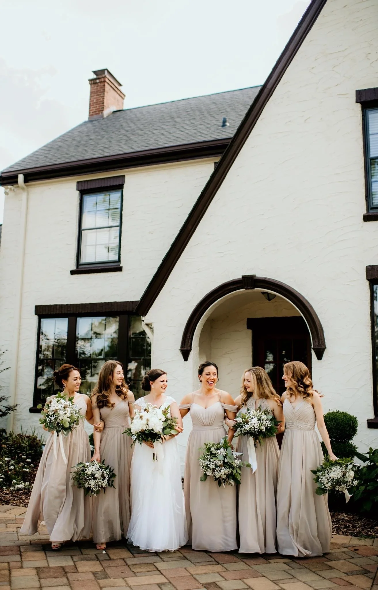 Group of bridesmaids in beige dresses standing outdoors in front of a large house, holding bouquets of white flowers. They are smiling and talking with each other, with the bride in the center.