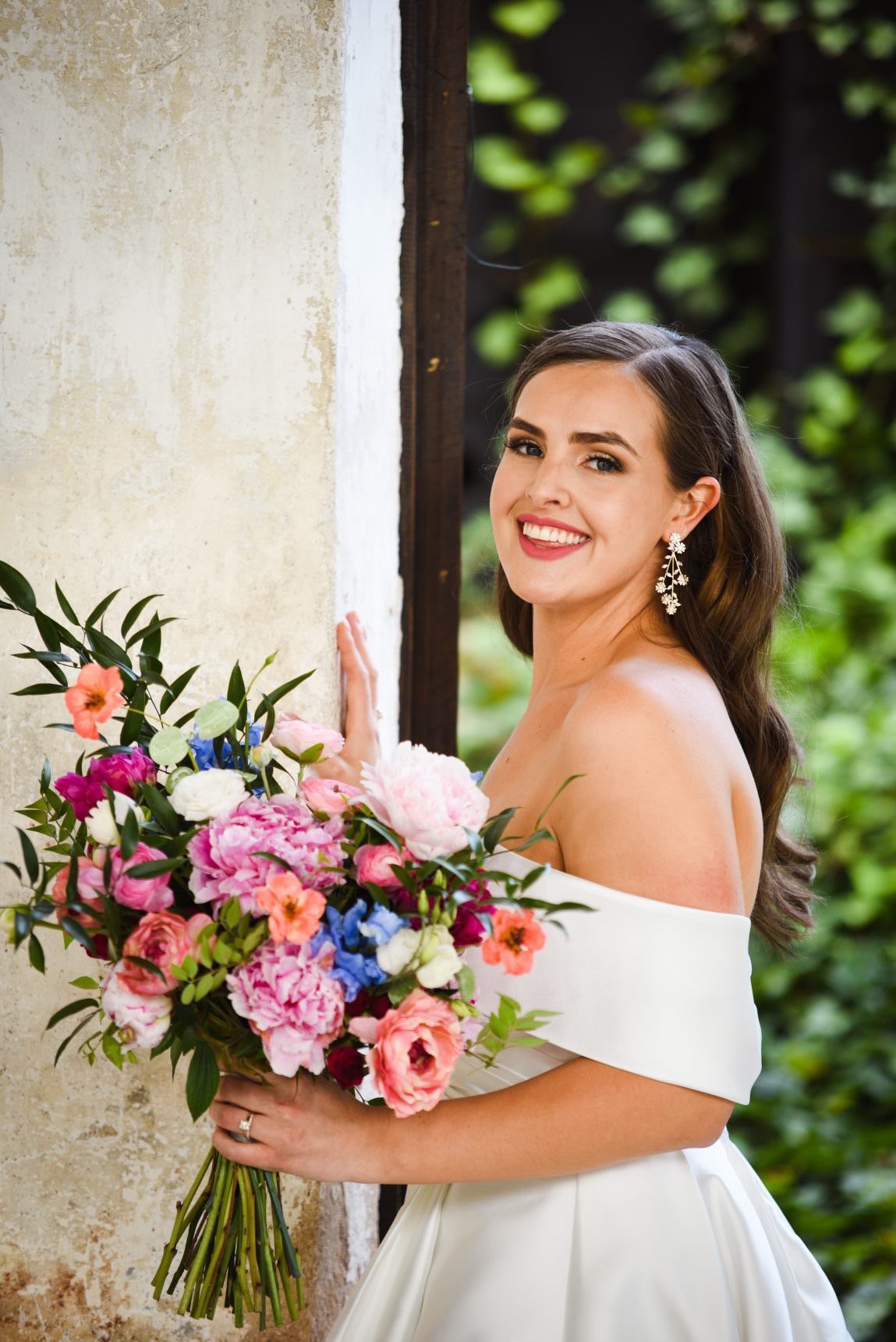 A woman in a white off-the-shoulder dress holding a large colorful bouquet of flowers. She is smiling and wearing elegant earrings, standing outdoors next to a stone wall with greenery in the background.