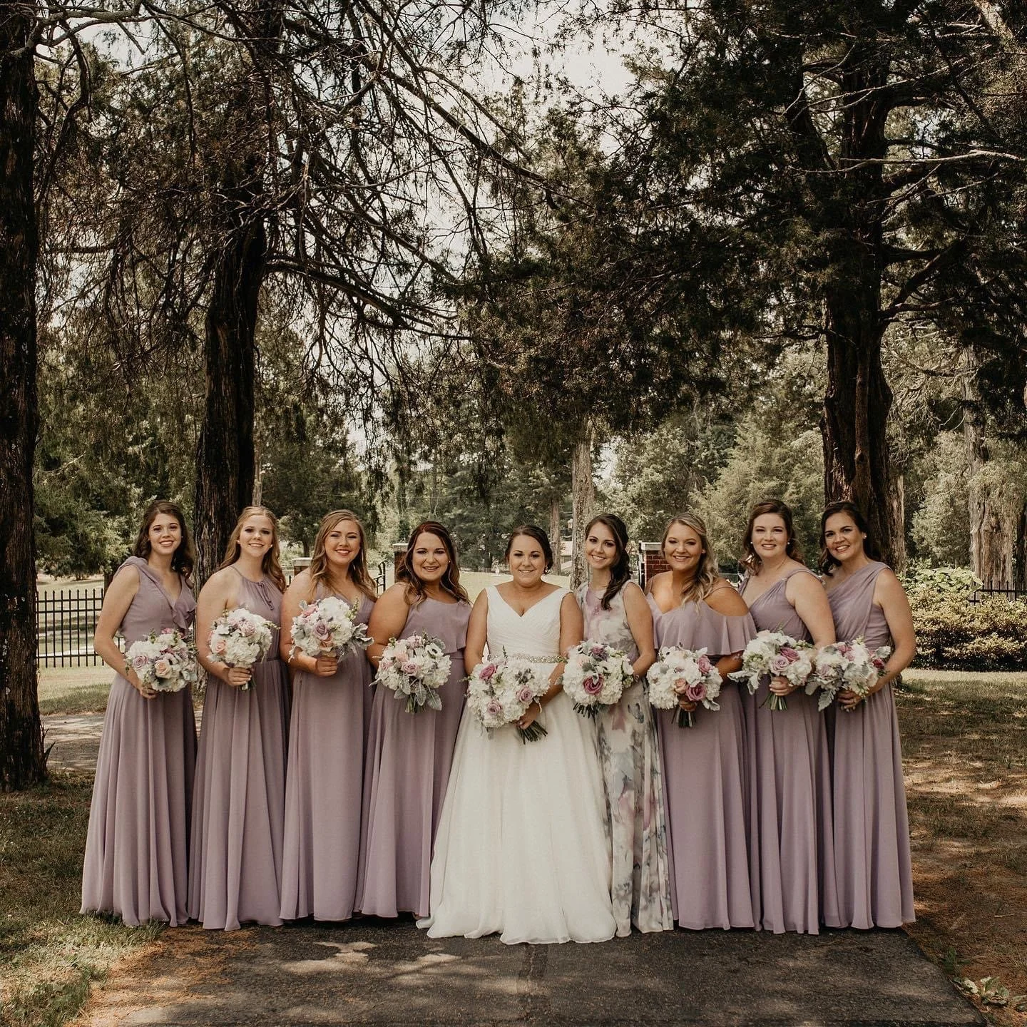 A group of nine women, including a bride in a white wedding dress and eight bridesmaids in lavender dresses, standing outdoors on a sunny day, holding bouquets of pink and white flowers and smiling.