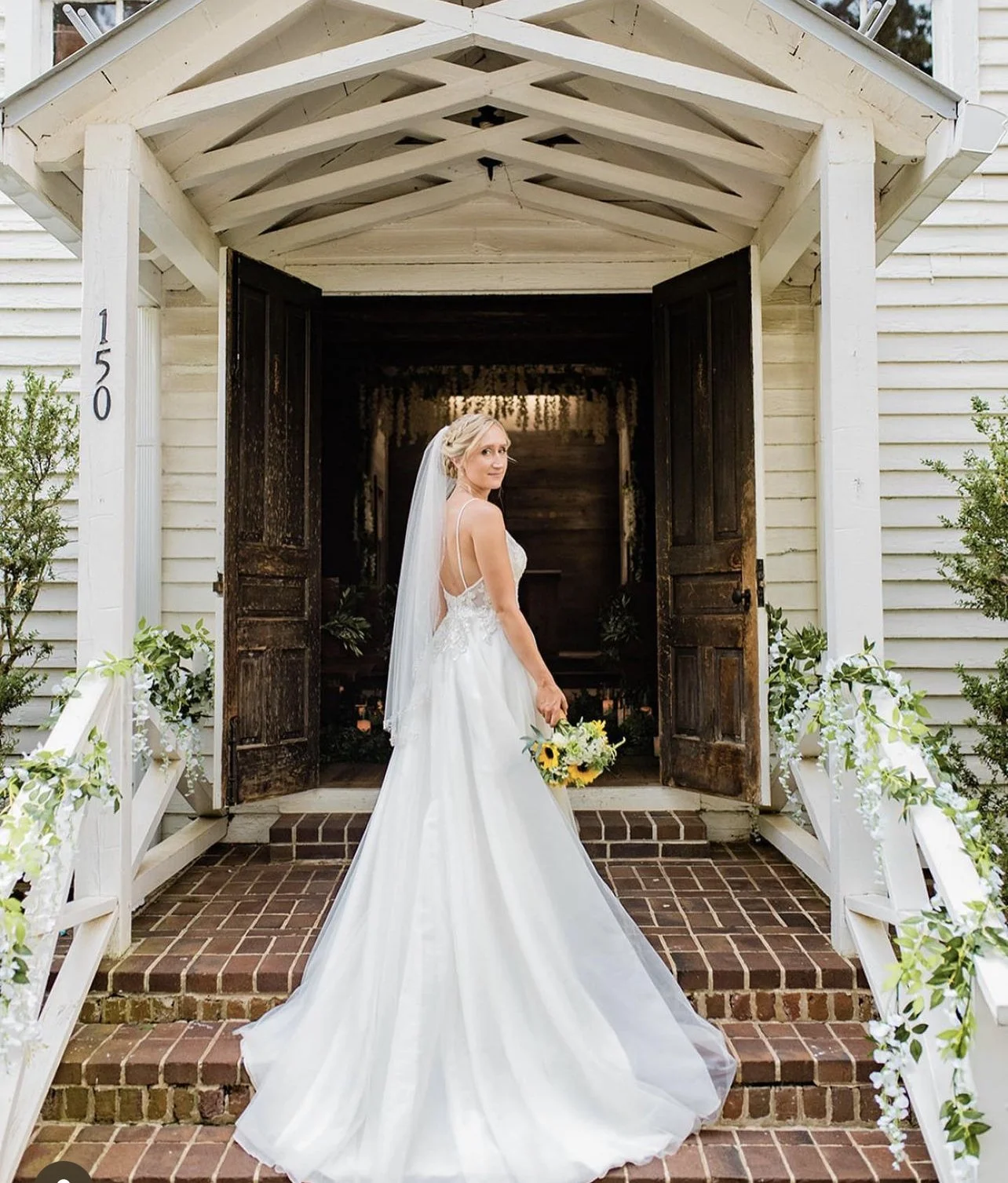 A bride in a white wedding dress holding a bouquet of sunflowers and greenery, standing on brick steps in front of a rustic wooden door at the entrance of a white house with floral decorations on the railing.