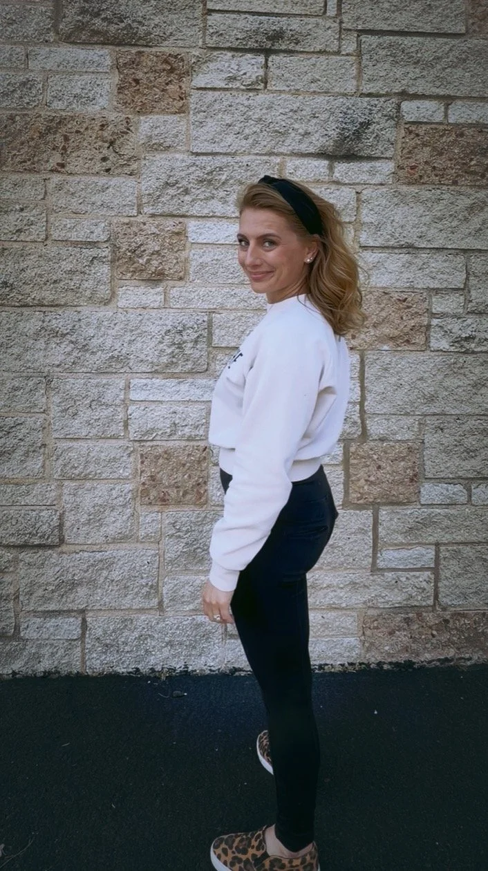 A woman with shoulder-length wavy hair wearing a black headband, white sweatshirt, black pants, and leopard-print shoes poses sideways against a textured stone wall, looking at the camera and smiling.