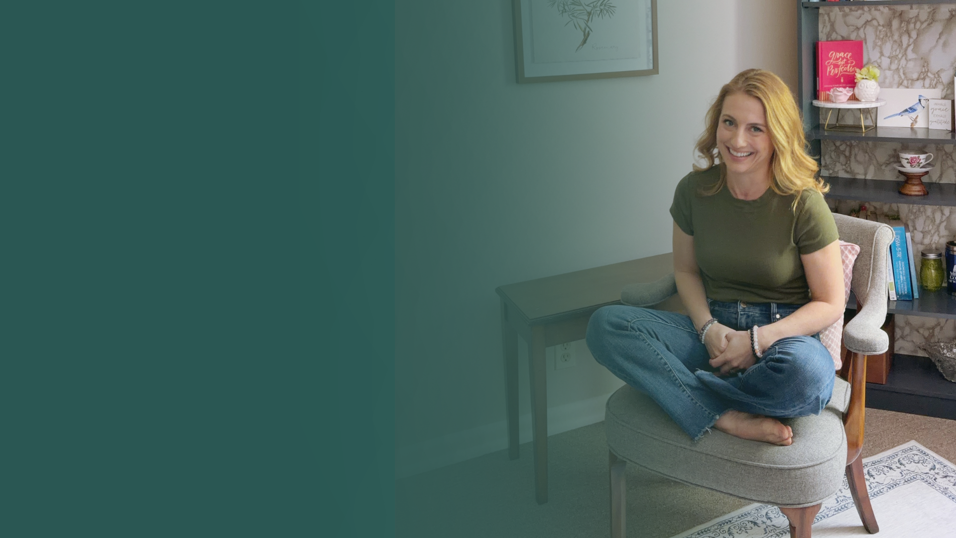 A woman with blonde wavy hair sitting cross-legged on a gray armchair in a cozy living room, smiling at the camera. She is wearing a green t-shirt and blue jeans, with jewelry on her wrists. Behind her, there's a patterned bookshelf with decorative items, books, and artwork on the wall.