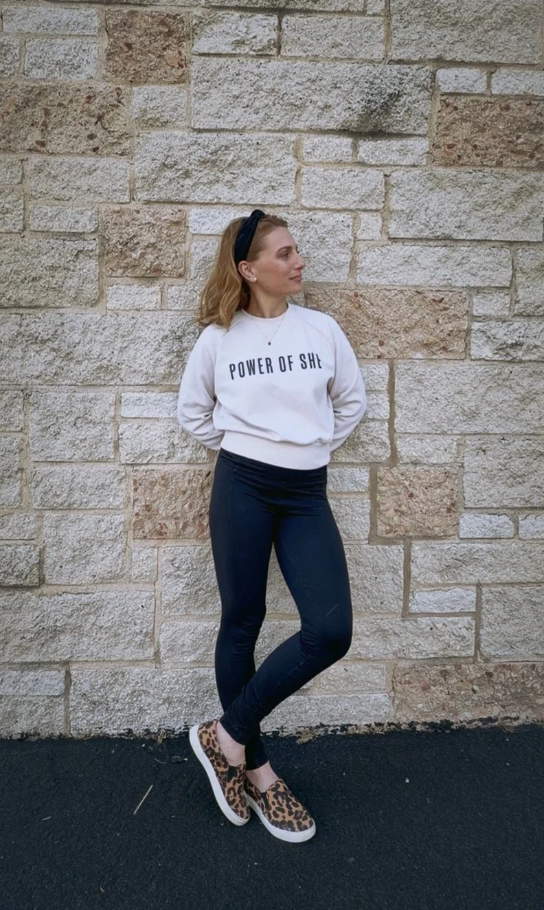 A woman with blonde hair wearing a white sweatshirt with 'Power of She' written on it, black high-waisted pants, leopard print slip-on shoes, and a black headband, standing against a brick wall.