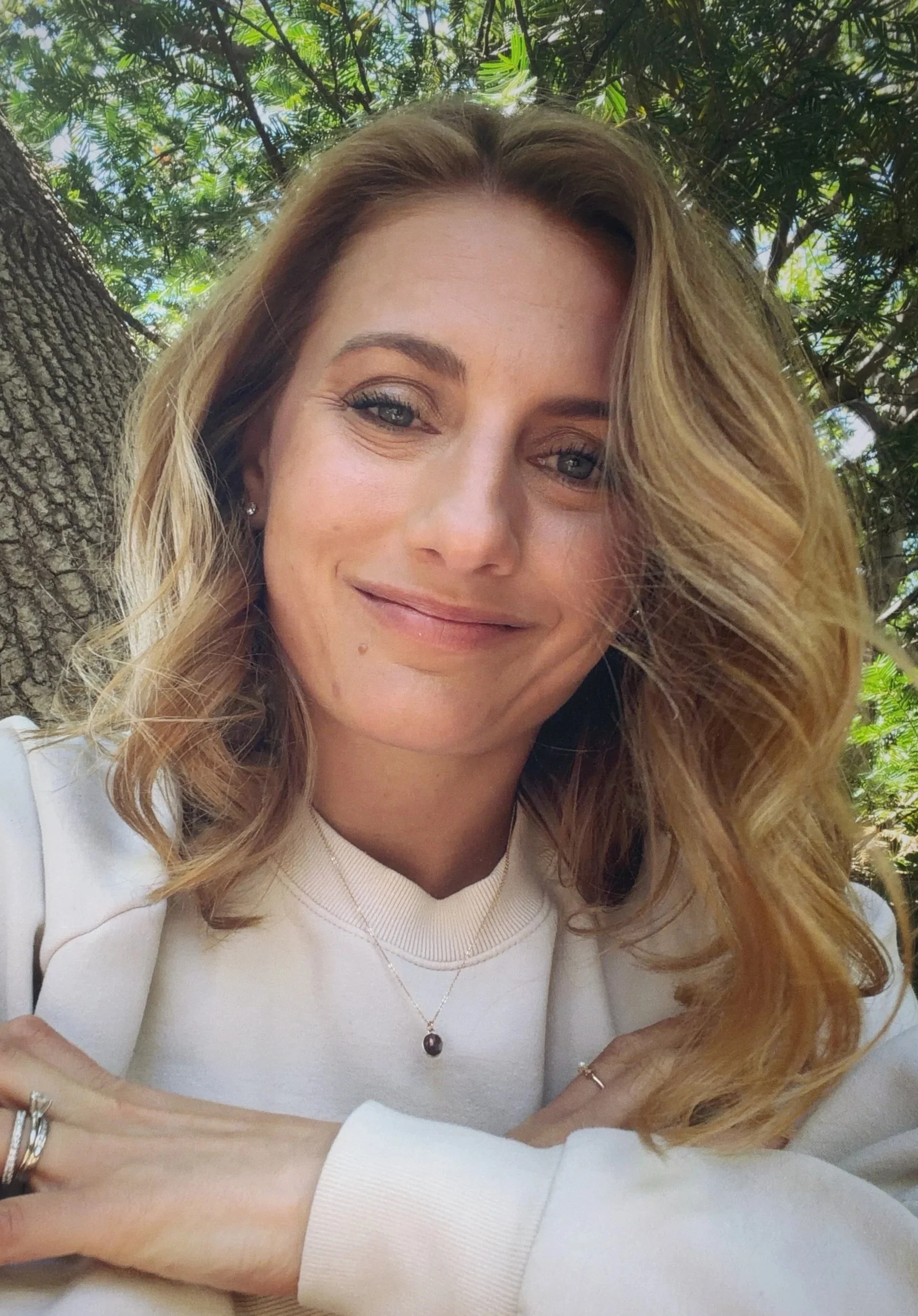 A woman with light brown, wavy hair, wearing makeup, a white long sleeve shirt, a necklace, and jewelry on her wrists, sitting outdoors with green tree foliage in the background, smiling and looking at the camera.