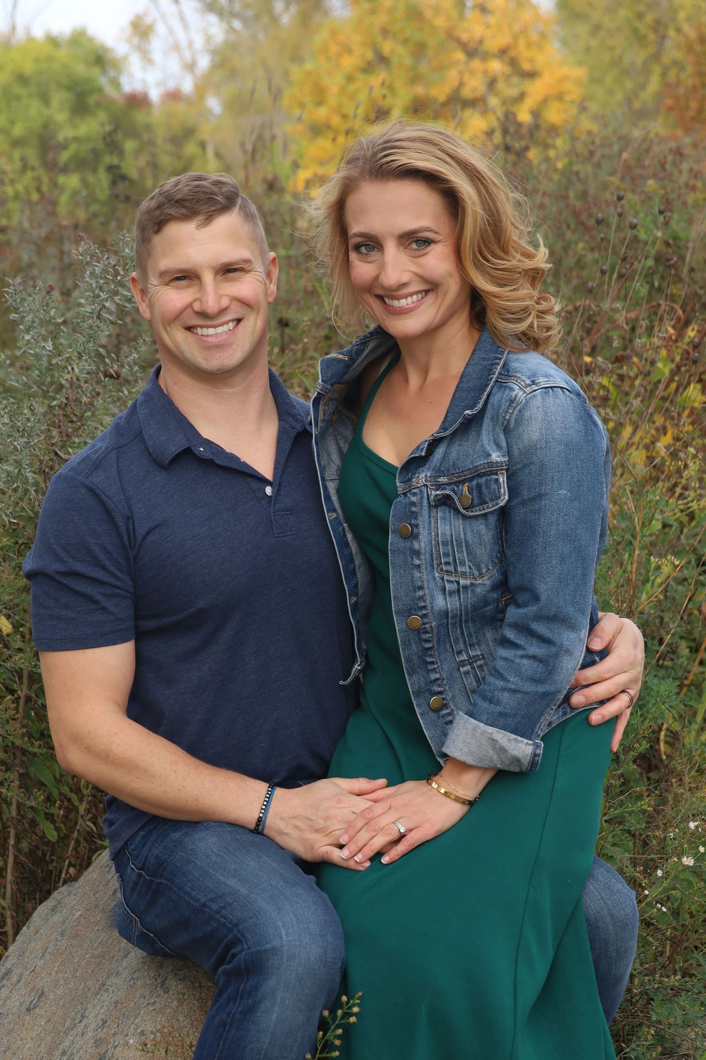 A smiling couple sitting on a large rock outdoors in a natural setting with trees showing fall foliage. The woman is wearing a denim jacket and green dress, and the man is in a navy shirt and jeans.