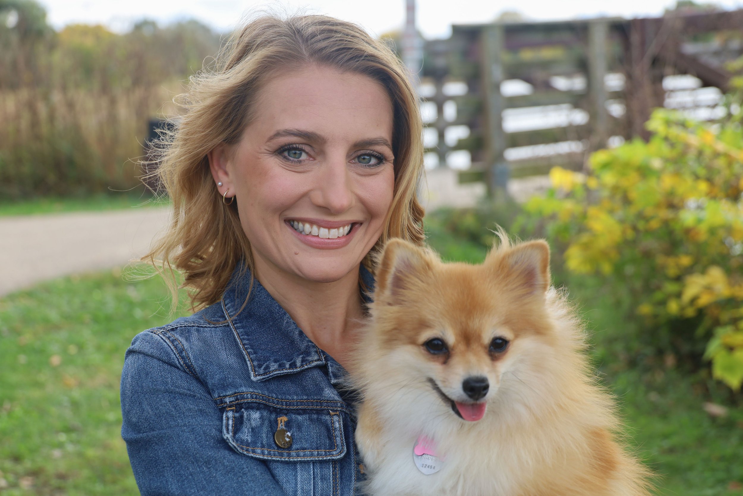 A woman smiling and holding a fluffy dog outdoors in a park-like setting with trees and a wooden fence in the background.