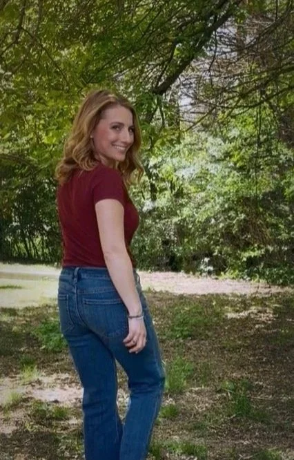 A woman with shoulder-length red hair, wearing a maroon t-shirt and blue jeans, standing outdoors in a green, wooded area with sunlight filtering through the trees.