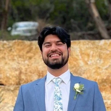 Man in a blue suit smiling outdoors with a background of trees and a stone wall.