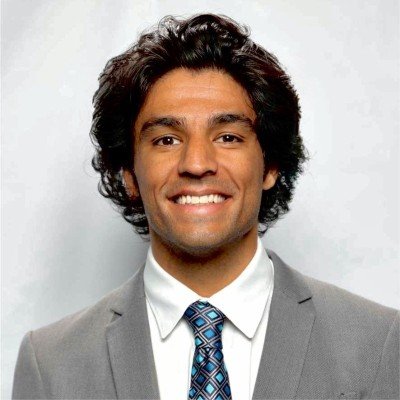 A man with curly black hair wearing a gray suit, white shirt, and patterned tie, smiling against a light gray background.