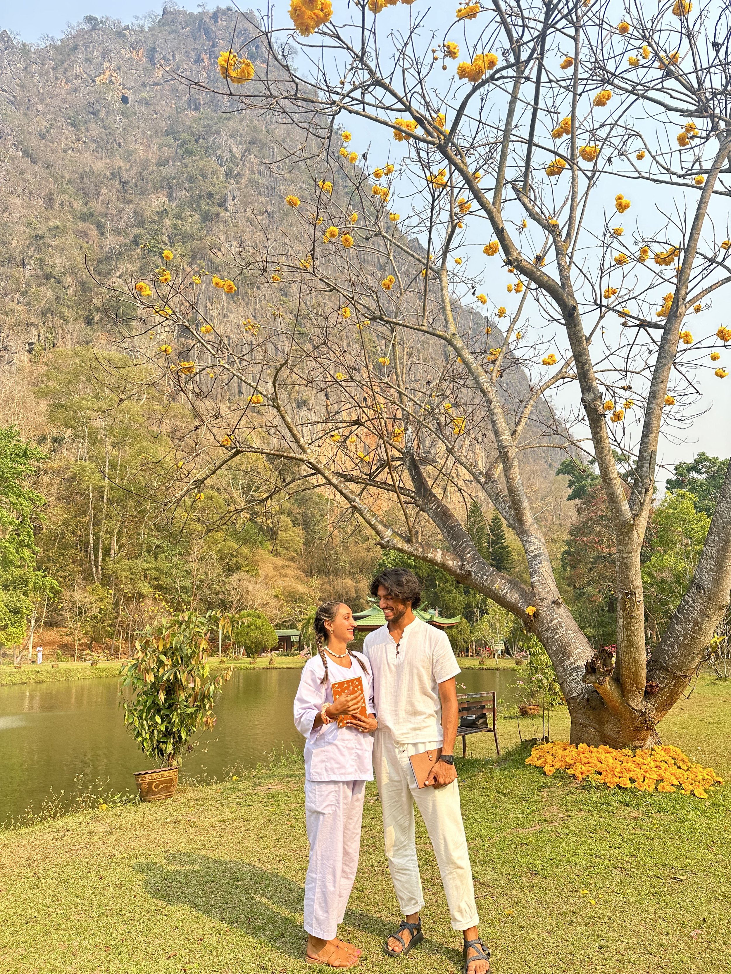 A man and a woman standing by a pond, smiling at each other, with a large tree with yellow flowers behind them, surrounded by a scenic landscape with mountains and more trees.