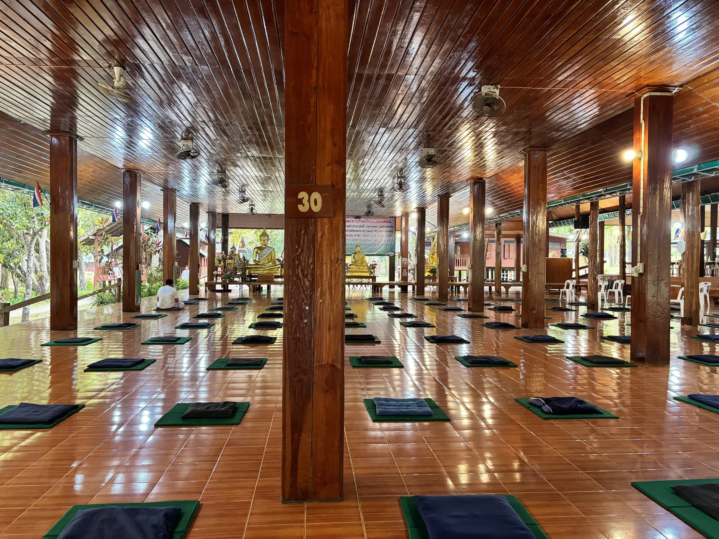 Indoor space with a wooden ceiling and columns, multiple meditation mats laid out on a shiny tiled floor, statues of Buddha at the front, and Thai flags hanging on the side.