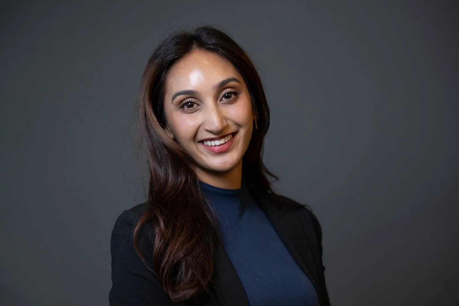 A woman with long dark hair smiling at the camera, wearing a black blazer and a dark blue top, standing against a plain dark gray background.