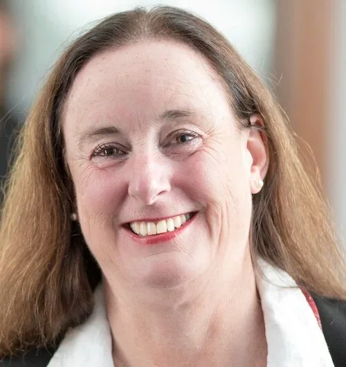 Close-up photo of a smiling woman with long, wavy brown hair, wearing a white blouse and small earrings.