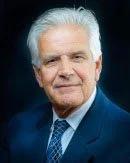 A senior man with white hair wearing a dark blue suit, light blue shirt, and patterned tie, posing against a dark background.