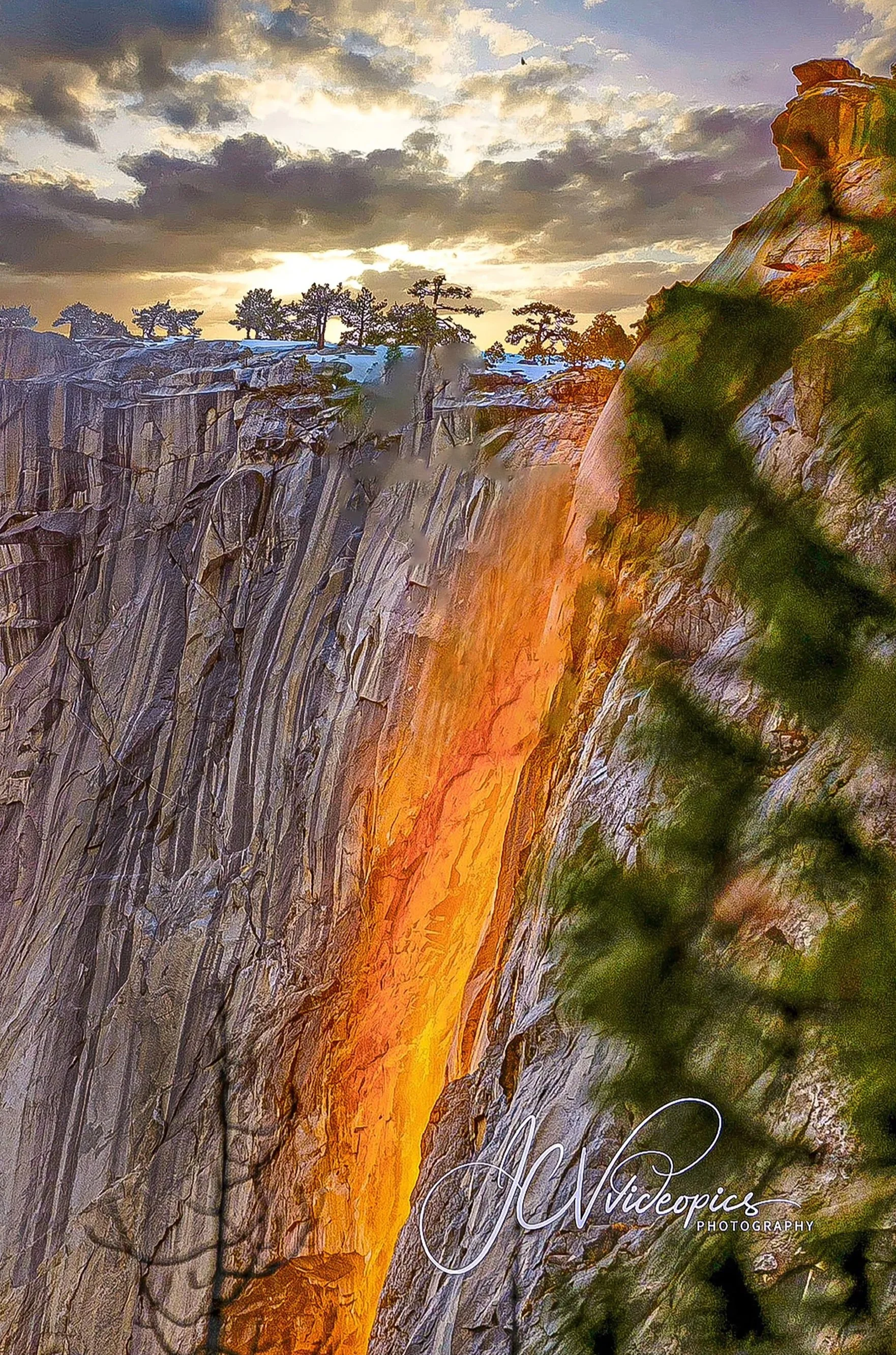 A waterfall flowing down a rocky cliff at sunset, with trees on top and dark clouds in the sky, and a signature at the bottom right corner reading 'Nicole Nicotine Photography'.