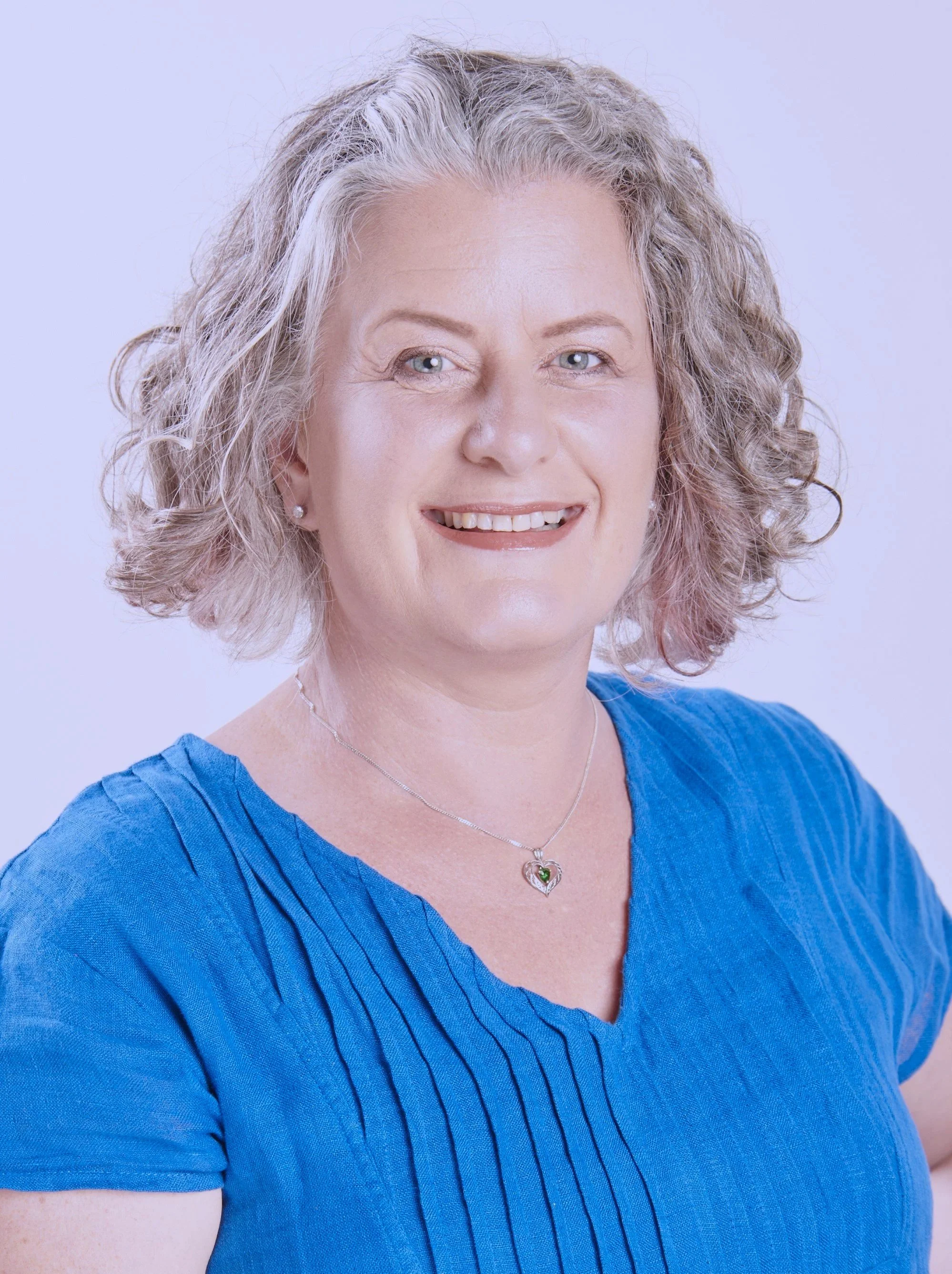 A middle-aged woman with curly gray hair and green eyes, smiling, wearing a blue top and a heart-shaped pendant necklace with a green stone, against a light background.