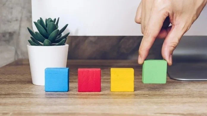 Colorful wooden blocks arranged in a row on a wooden surface with a potted succulent plant in the background, and a hand reaching to move the green block.