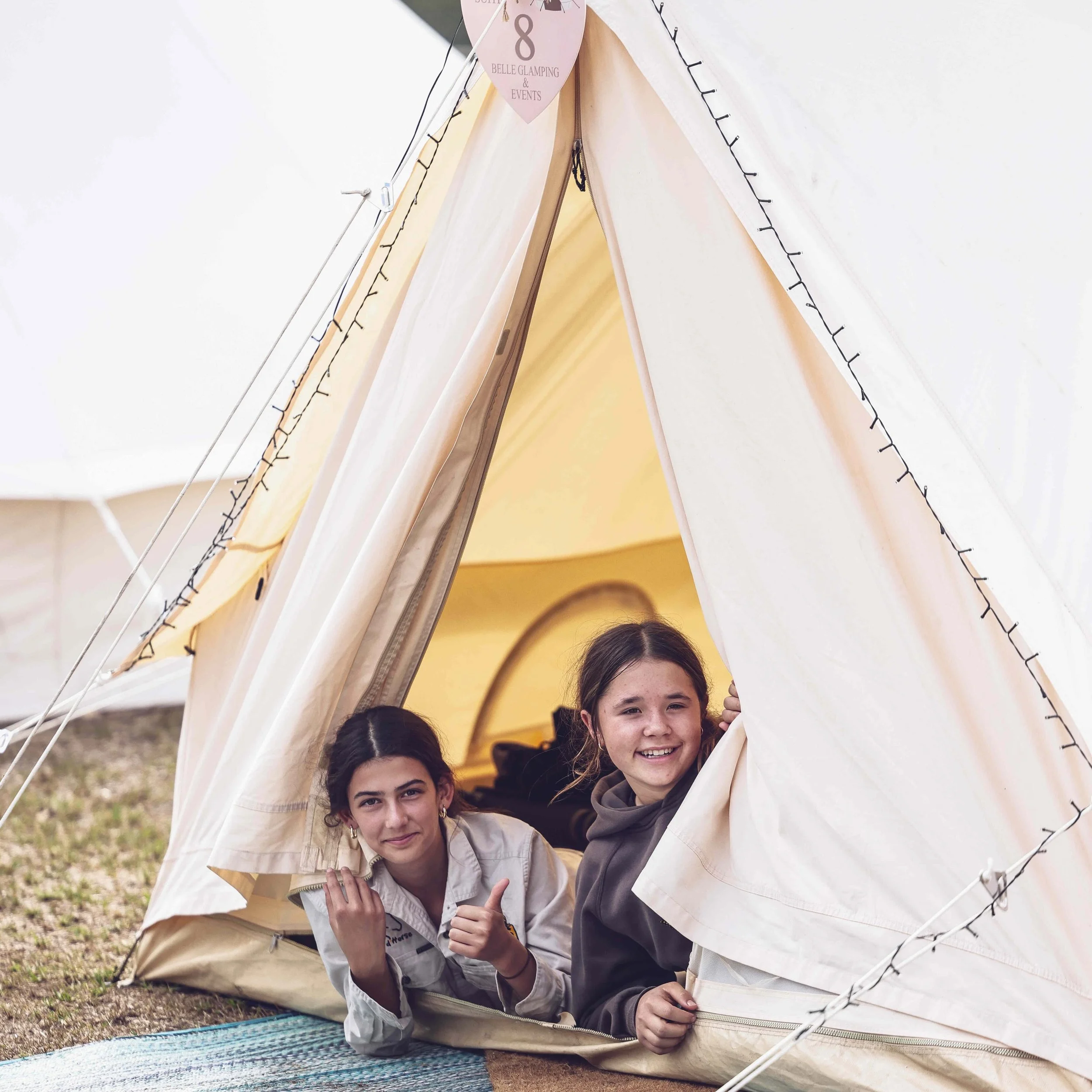 Three young girls smiling and peeking out from inside a small camping tent, outdoors on grass, with a blanket on the ground, during daytime.