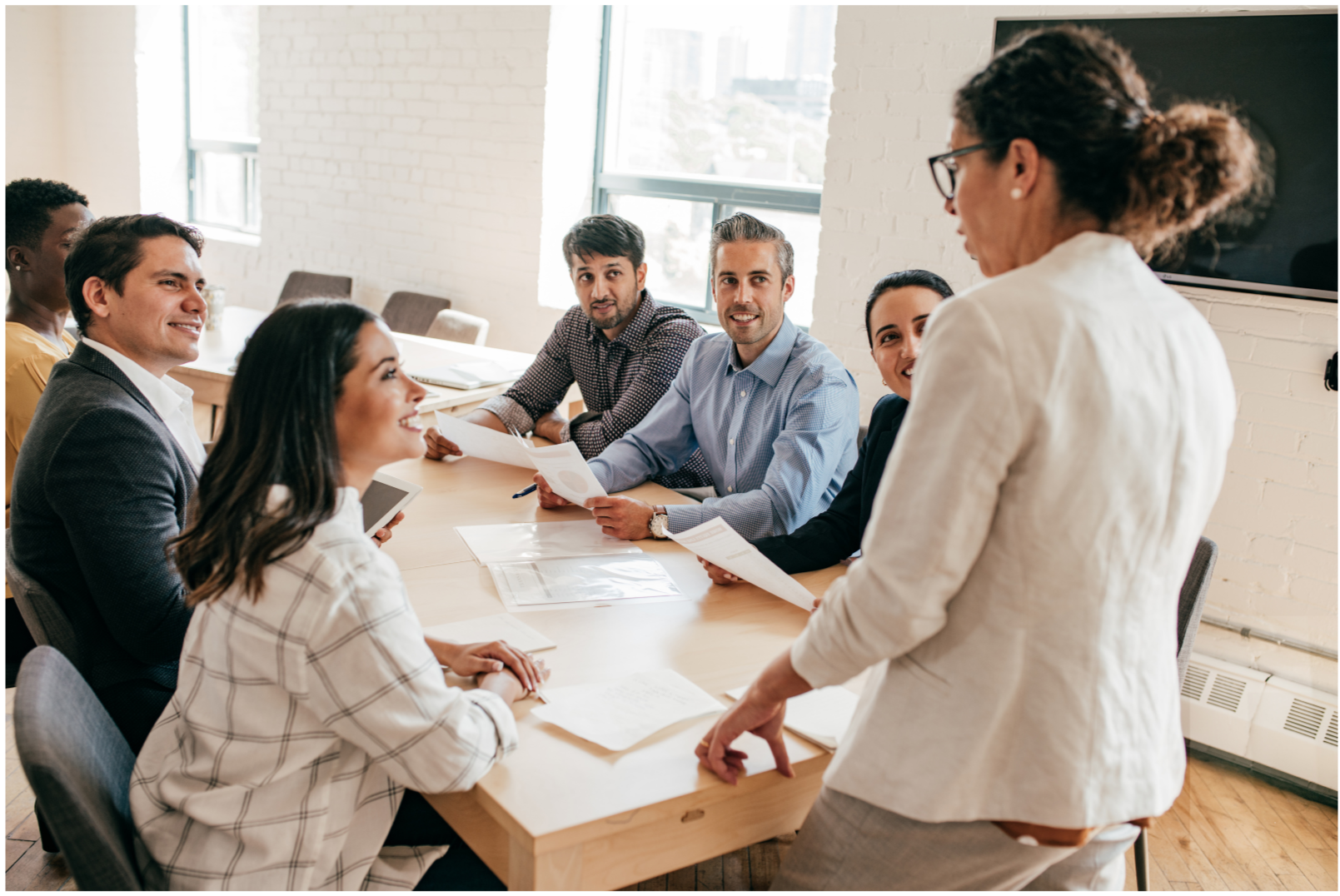 A group of business professionals sitting around a conference table listening to a woman standing and speaking.