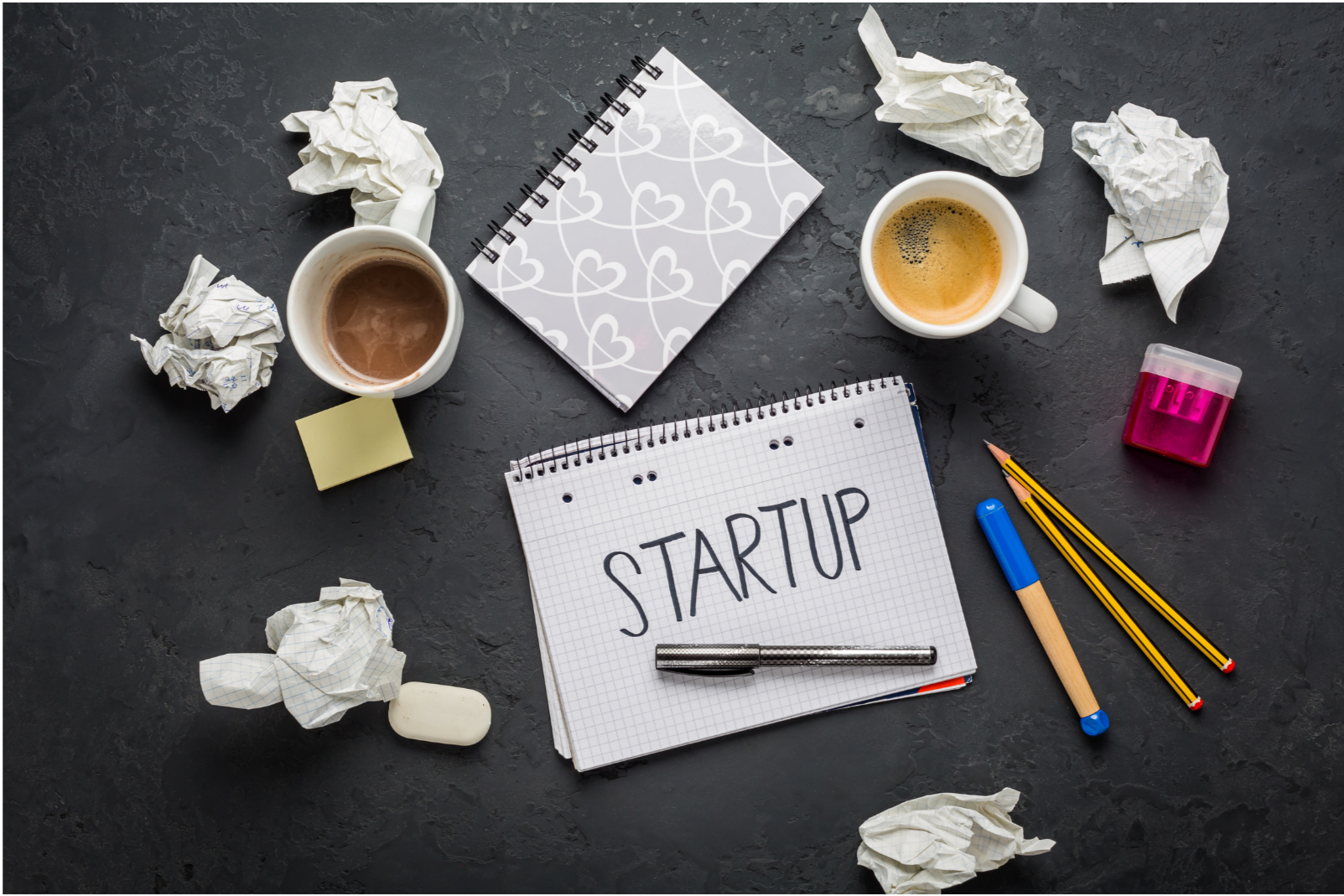Disorganized desk with crumpled paper, cups of coffee, a small notepad with a heart pattern, a graph paper notebook with 'STARTUP' written on it, a black and silver pen, two yellow pencils, a blue marker, pink sharpener, and sticky notes on a dark textured surface.
