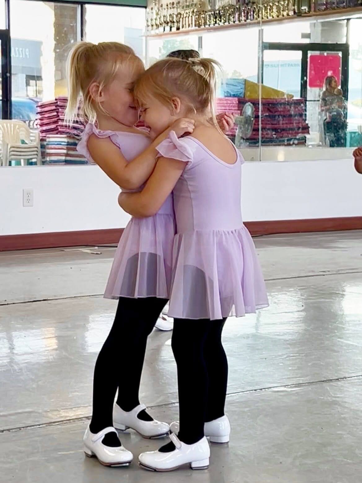 Two young girls with blonde hair in lavender dance dresses and black leggings sharing a close embrace and a hug during a dance class or event.