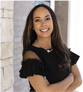 Woman with long dark hair smiling, wearing a black dress with ruffled sleeves and a necklace, standing indoors near a white brick wall.