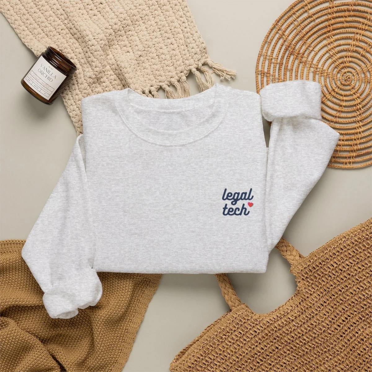 A white sweatshirt with 'legal tech' written on it, surrounded by woven beige and brown accessories and a brown candle labeled vanilla orchid.