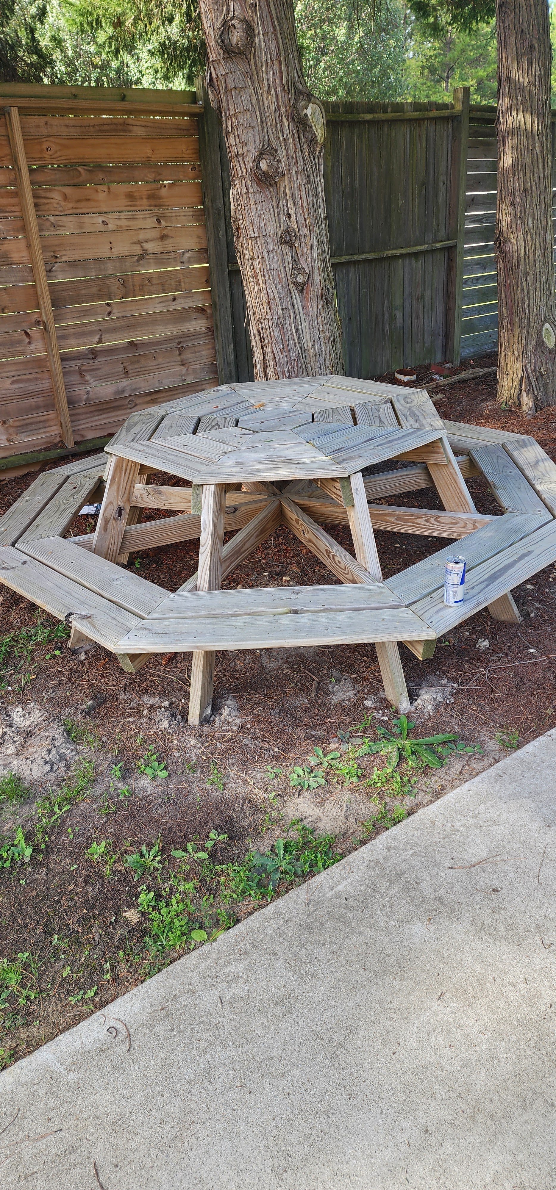 A wooden outdoor table and bench set with a geometric design, placed in a backyard with a dirt and grass ground, surrounded by trees and a wooden fence.