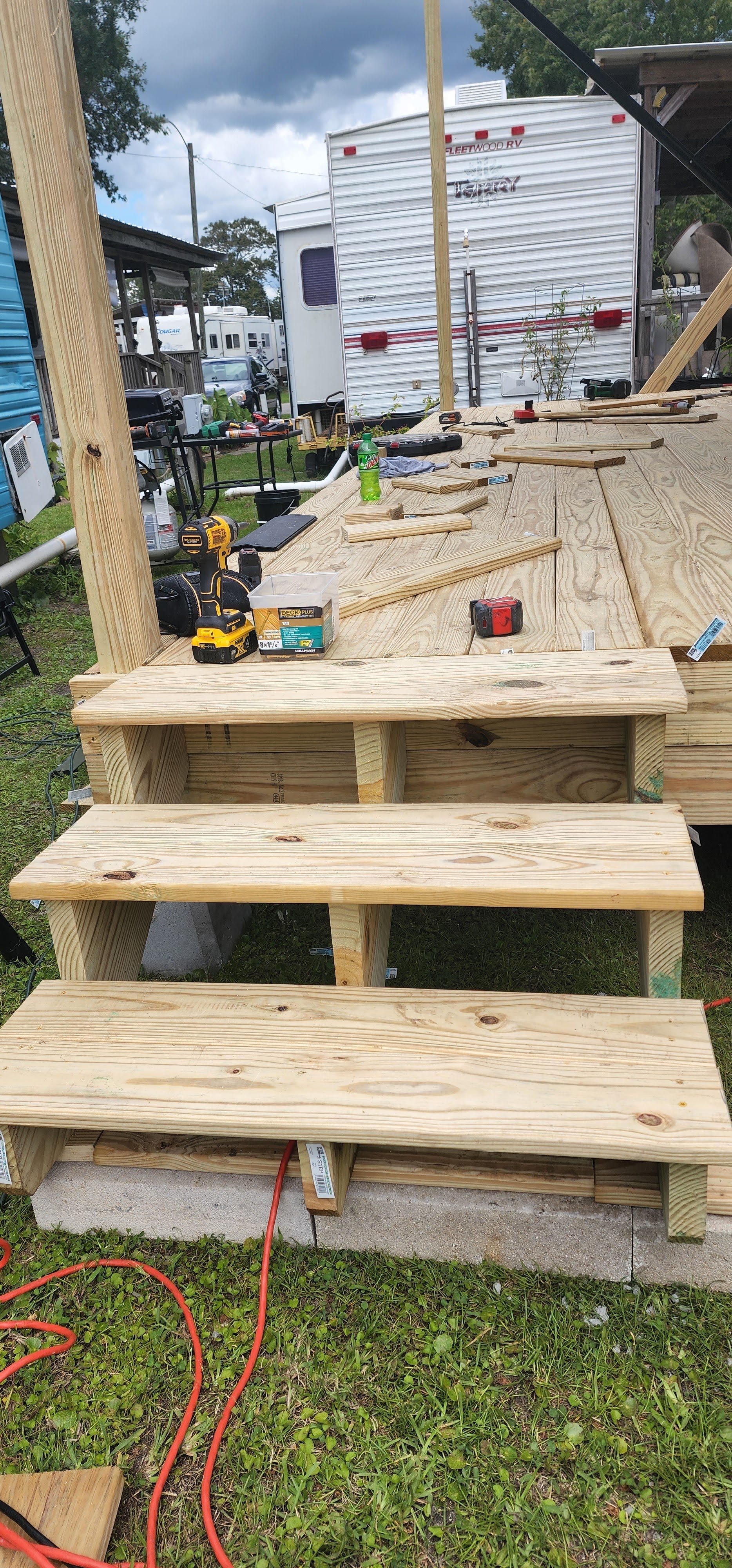 Construction of wooden staircase with plywood steps outdoors, surrounded by tools, a cordless drill, a soda bottle, and a workbench, with RVs and trees in the background.