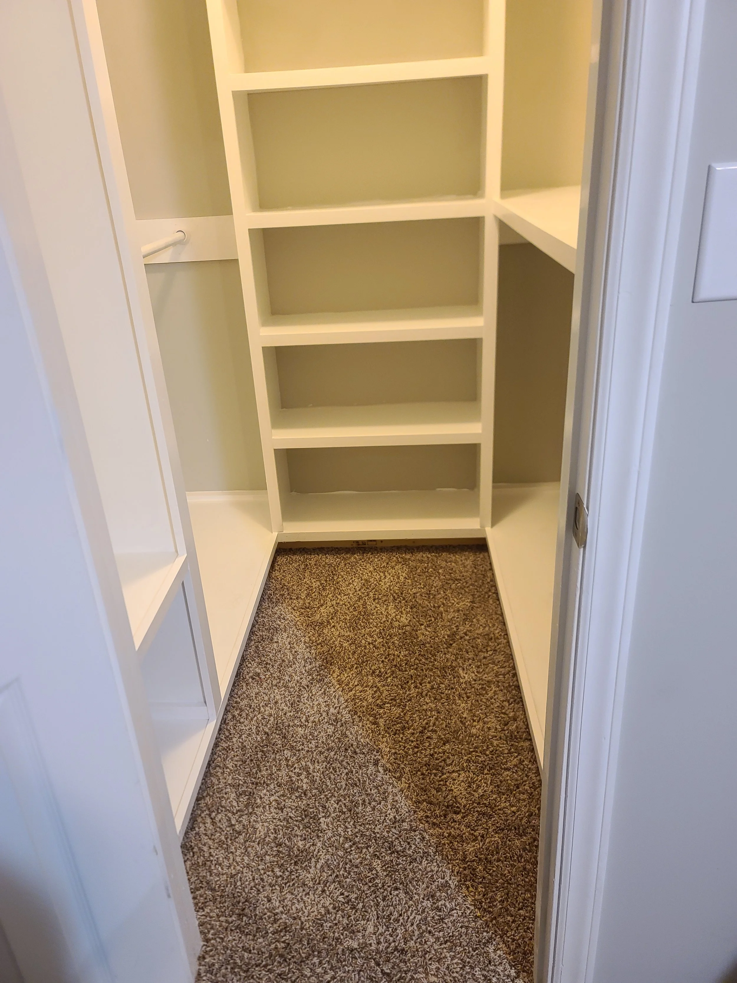 Empty walk-in closet with built-in white shelves on the left and right walls, beige walls, carpeted floor, and a small white shelf on the right side.