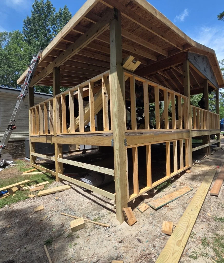 A two-story wooden deck under construction, with framing for the railings and supports visible. Construction tools and materials are scattered around, and a ladder leans against the structure.