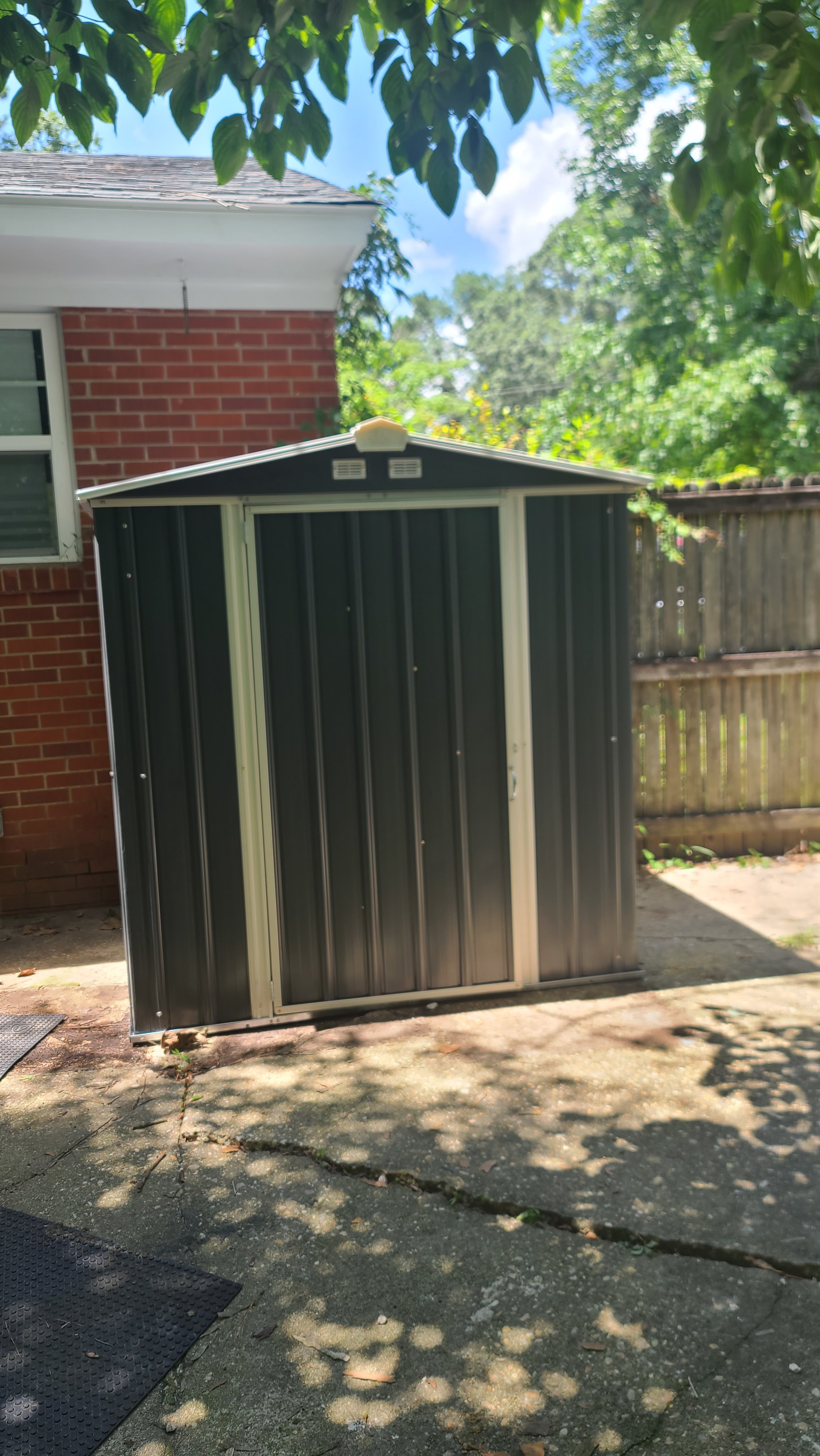 A small black metal shed with white framing is positioned on a concrete patio, situated next to a brick house with a window. There is a wooden fence to the right, and leafy trees overhead cast shadows on the ground, with a mostly clear blue sky in the background.