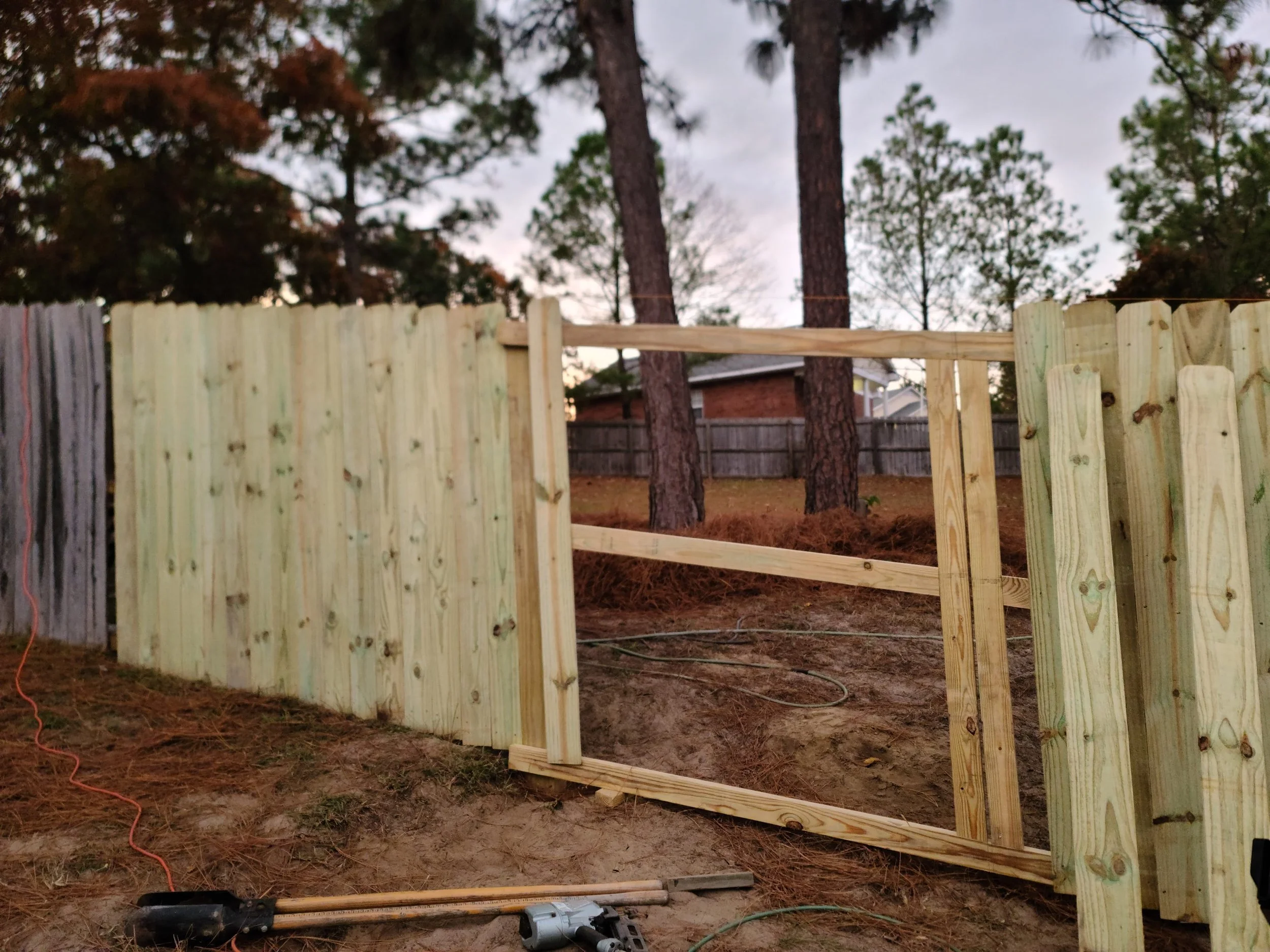 A backyard with a wooden fence in construction. The fence is partially built with vertical wooden planks and some horizontal support beams. There are trees in the background and tools on the ground, including a cordless drill and an extension cord.