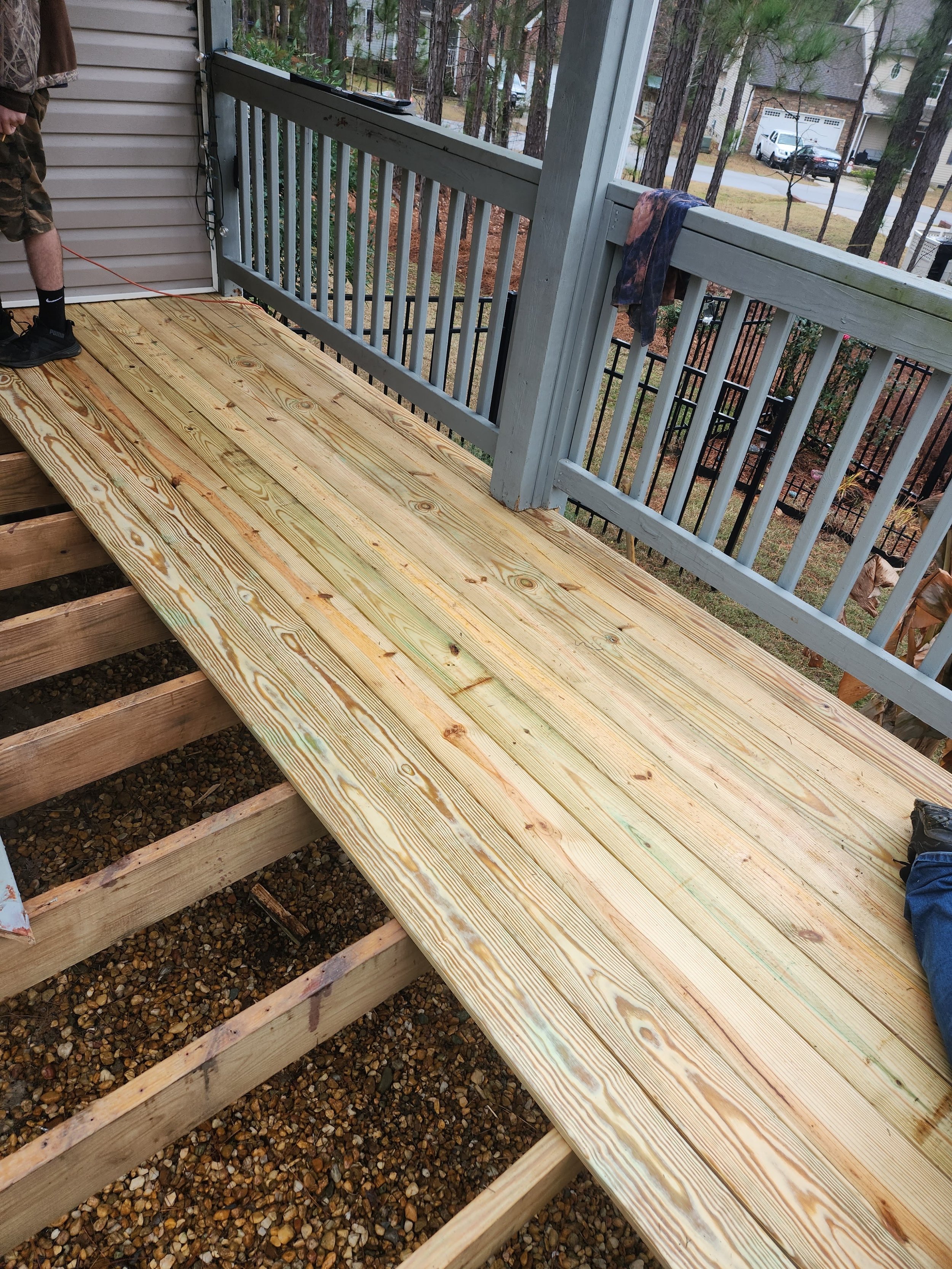 A photo of a porch railing with a wooden deck being built on a house's balcony, with visible stairs and surrounding trees in a residential neighborhood.