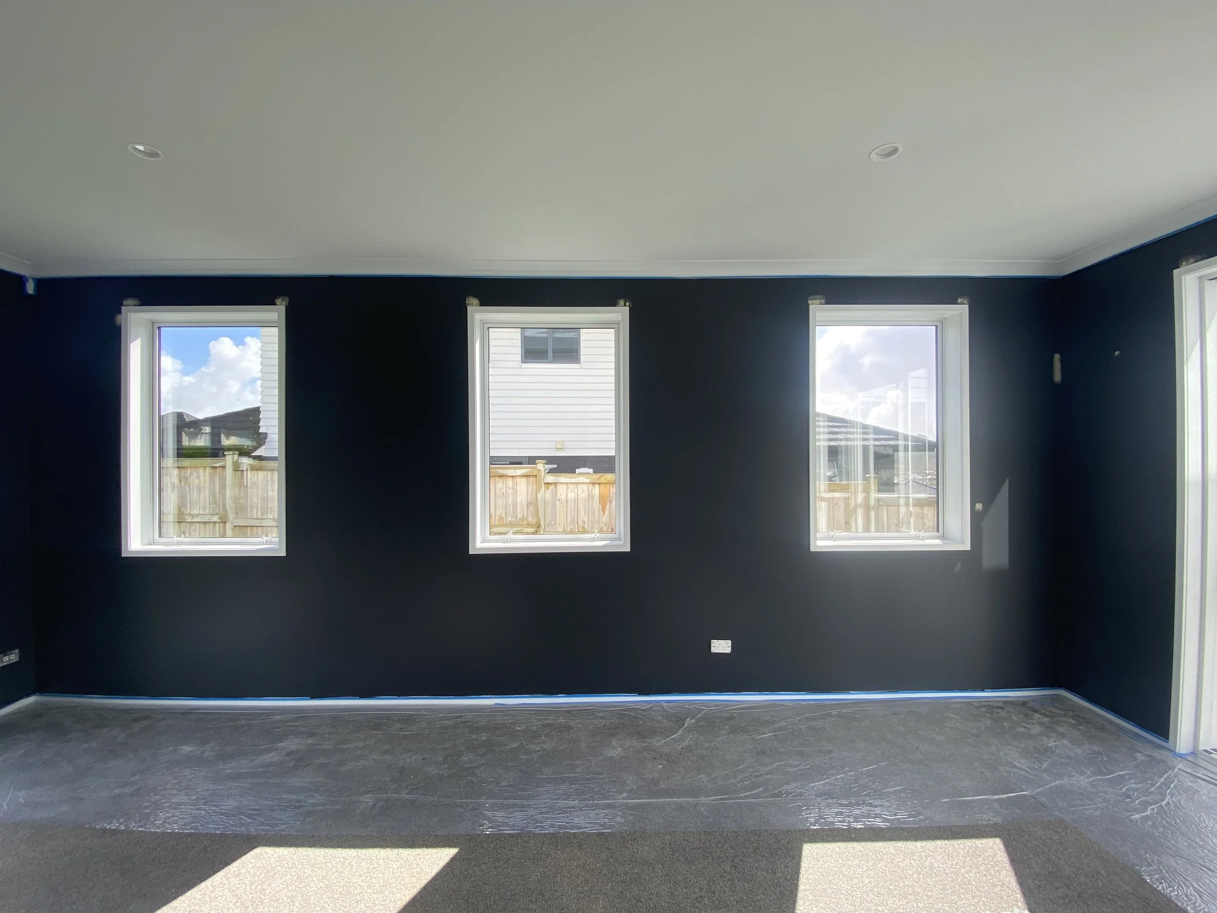 Interior of a room with black painted walls, three windows showing a partly cloudy sky and neighboring houses, and a dark floor.