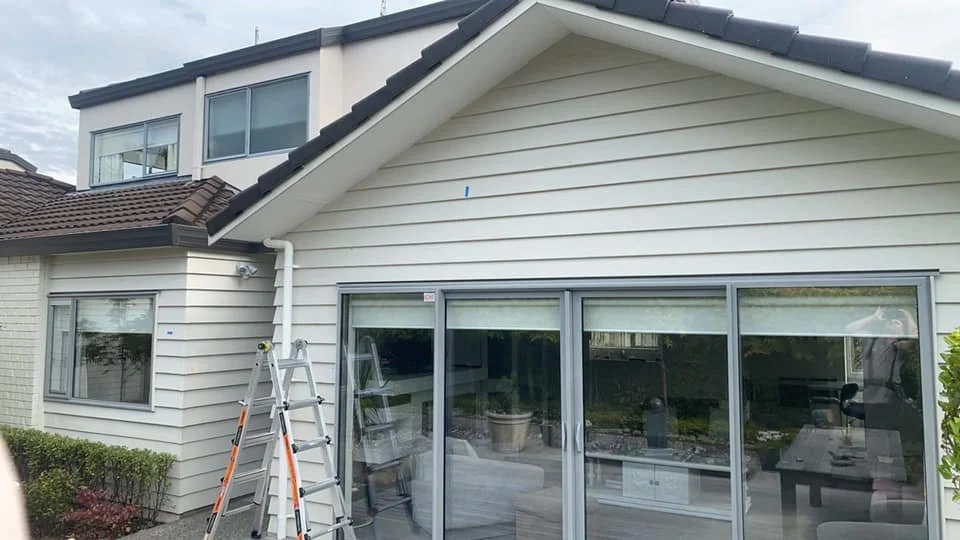 The back of a two-story house with beige siding, a sliding glass door, a ladder, and a hedge in the garden.