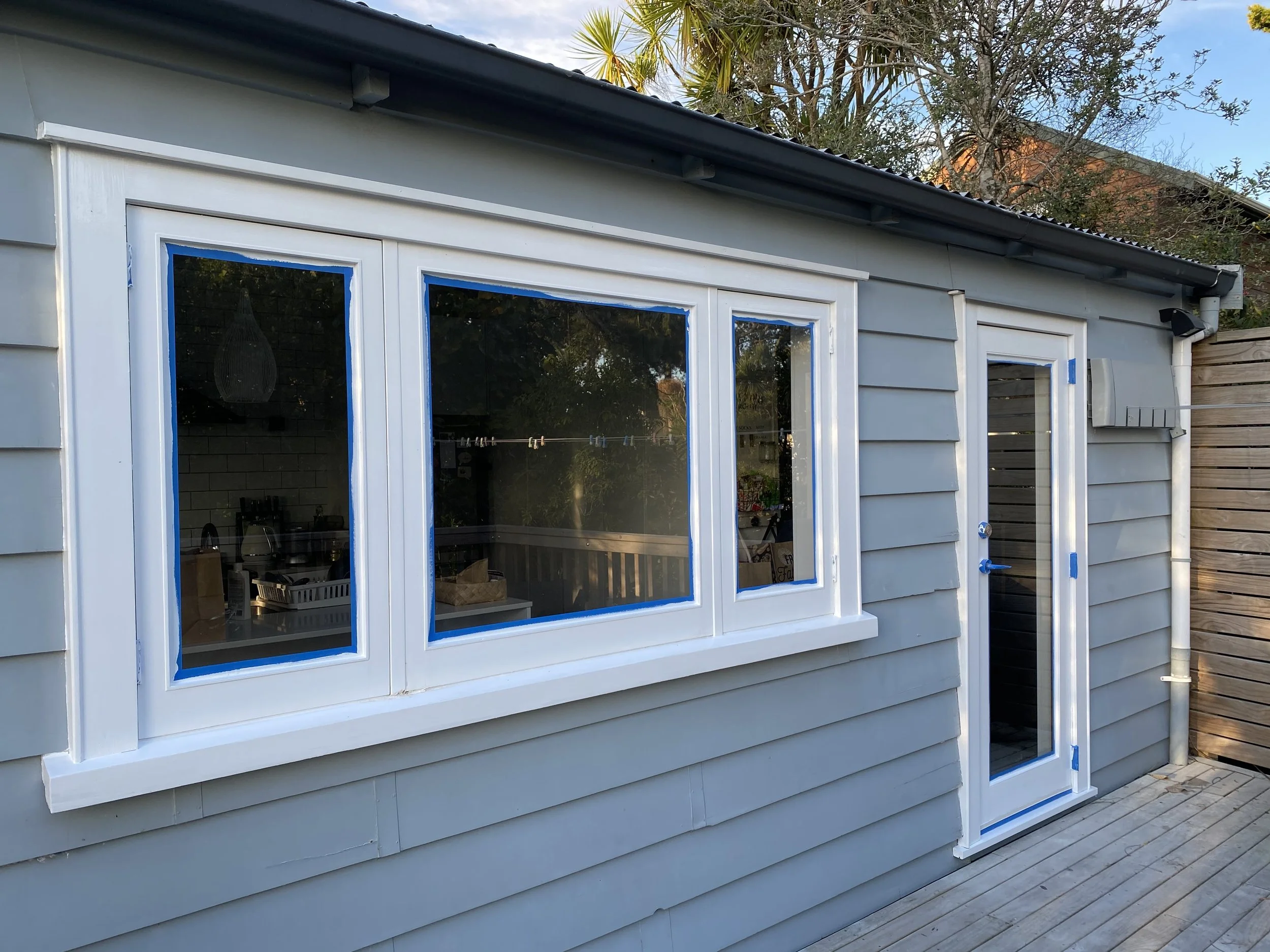 A house exterior with large glass windows framed with blue painter's tape, indicating painting or renovation work.