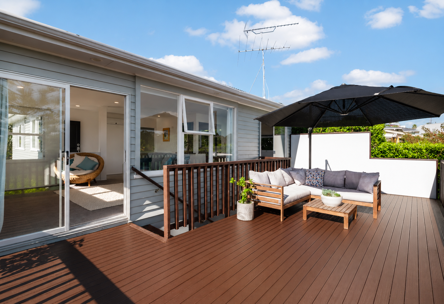 Outdoor balcony with a large seating area under a black umbrella, wooden railing, potted plants, and houses nearby under a blue sky.